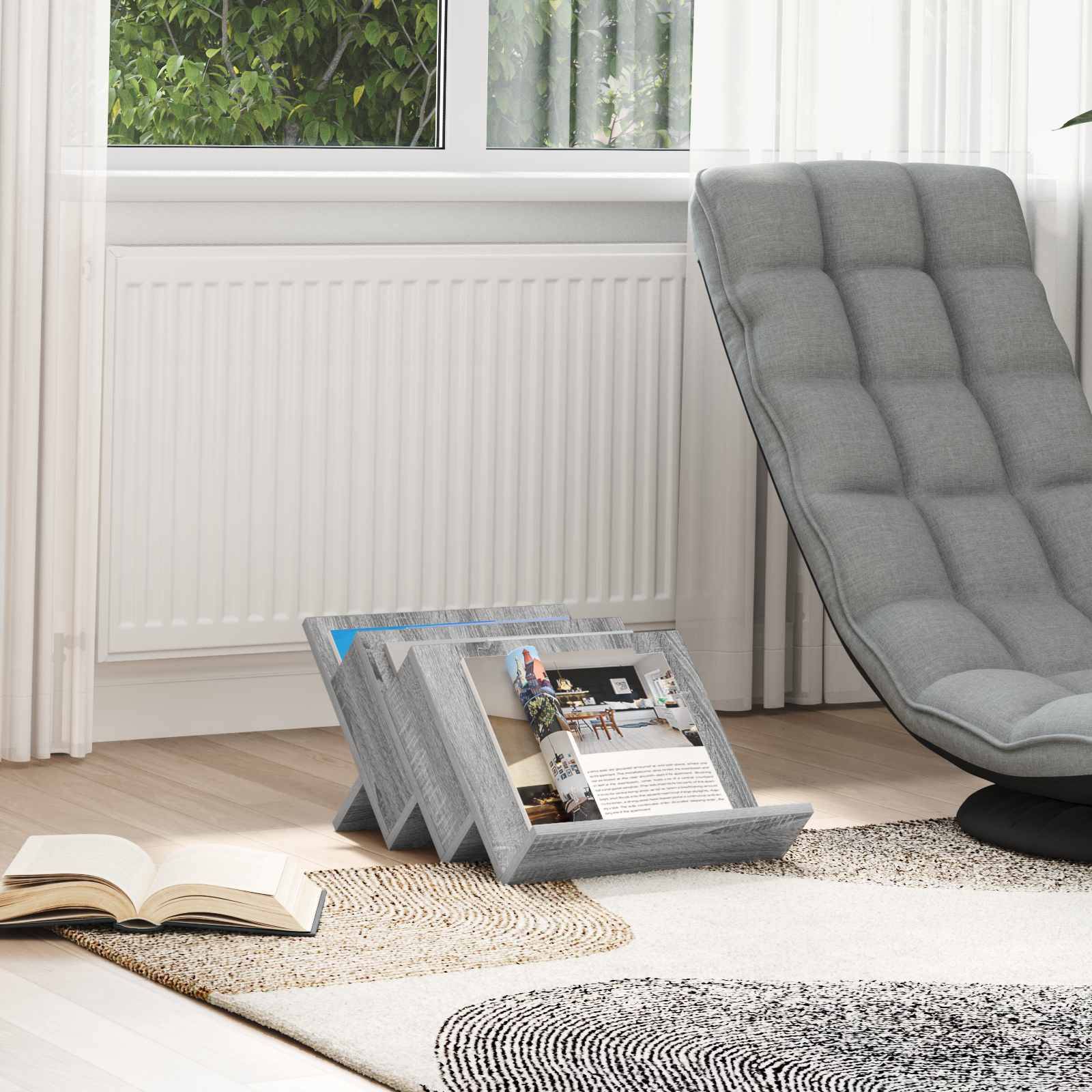 Ein gemütliches Zimmer mit einem grauen Sessel, einem aufgeschlagenen Buch und schräg auf dem Boden liegenden Zeitschriften in der Nähe eines großen Fensters, durch das Grünpflanzen zu sehen sind.