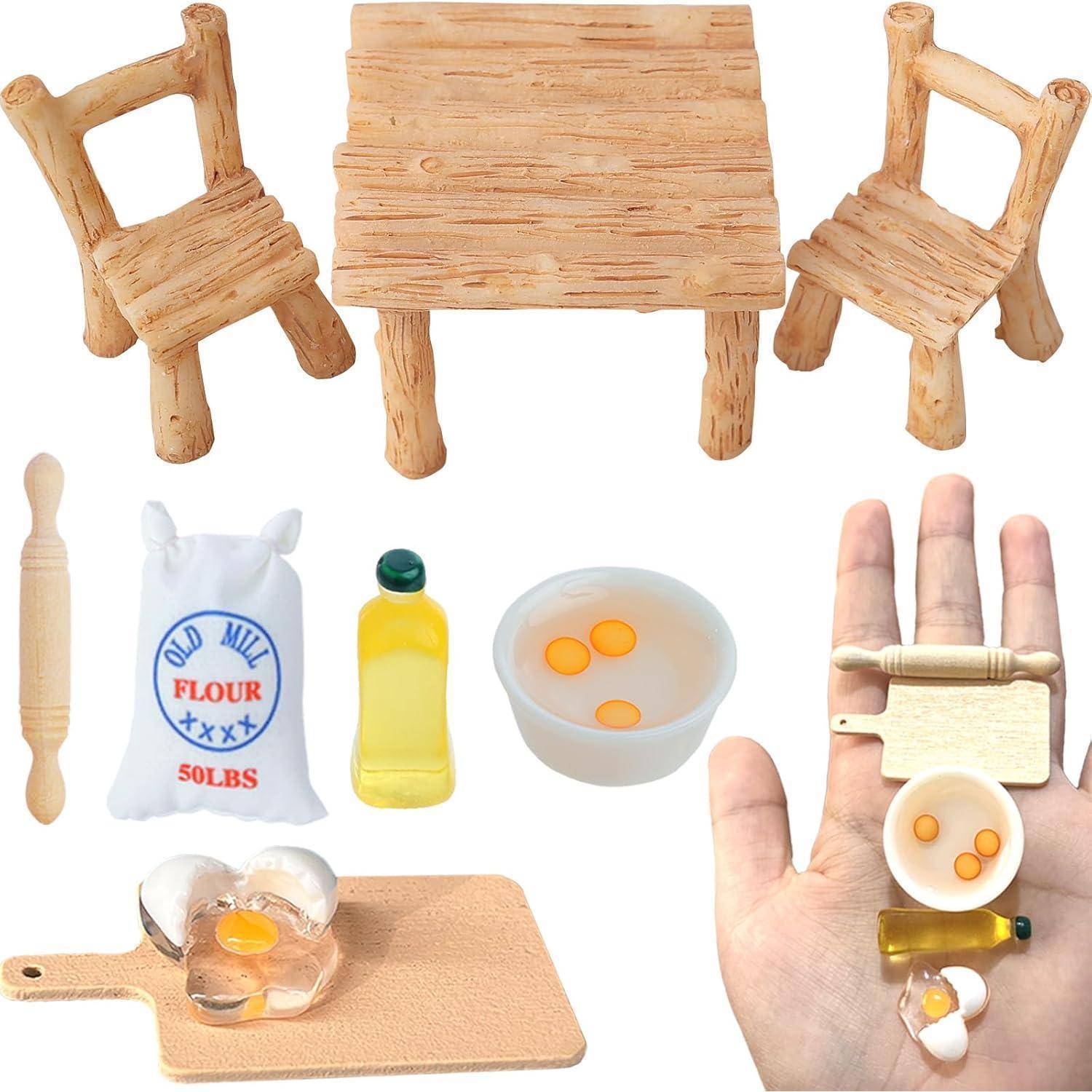 Miniatur-Küchenaufbau mit Holztisch und -stühlen, winzigem Küchengeschirr und Mini-Lebensmitteln wie Eiern, Öl und Mehl, die auf einer Hand gezeigt werden.