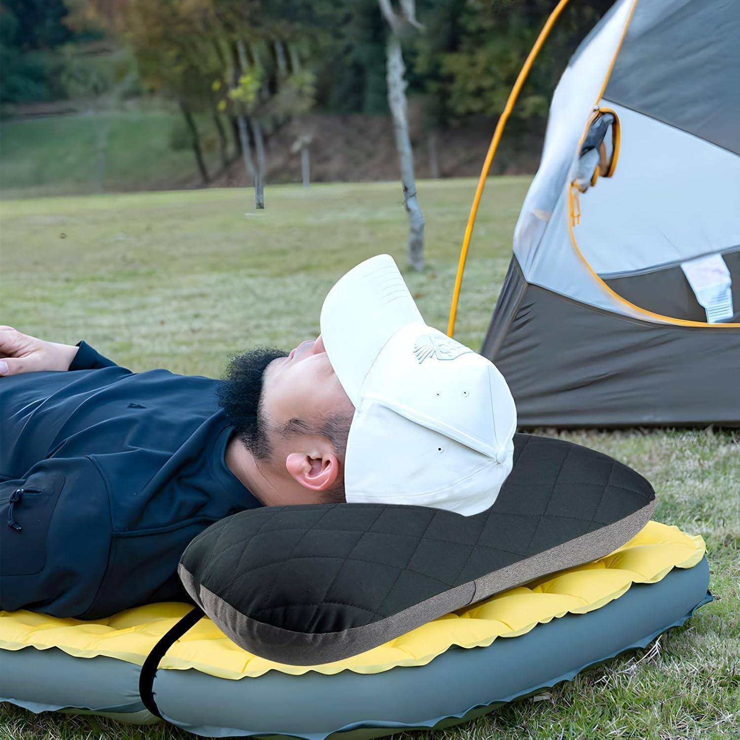 Person auf einer aufblasbaren Campingmatratze im Freien, trägt eine weiße Kappe, in der Nähe eines Zeltes in einem Grasbereich.