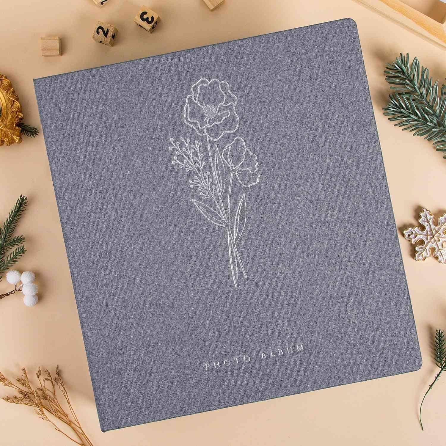 Ein graues Fotoalbum mit einem Blumenmuster auf dem Einband ist umgeben von Weihnachtsdekorationen wie Tannenzweigen und goldenen Christbaumkugeln.