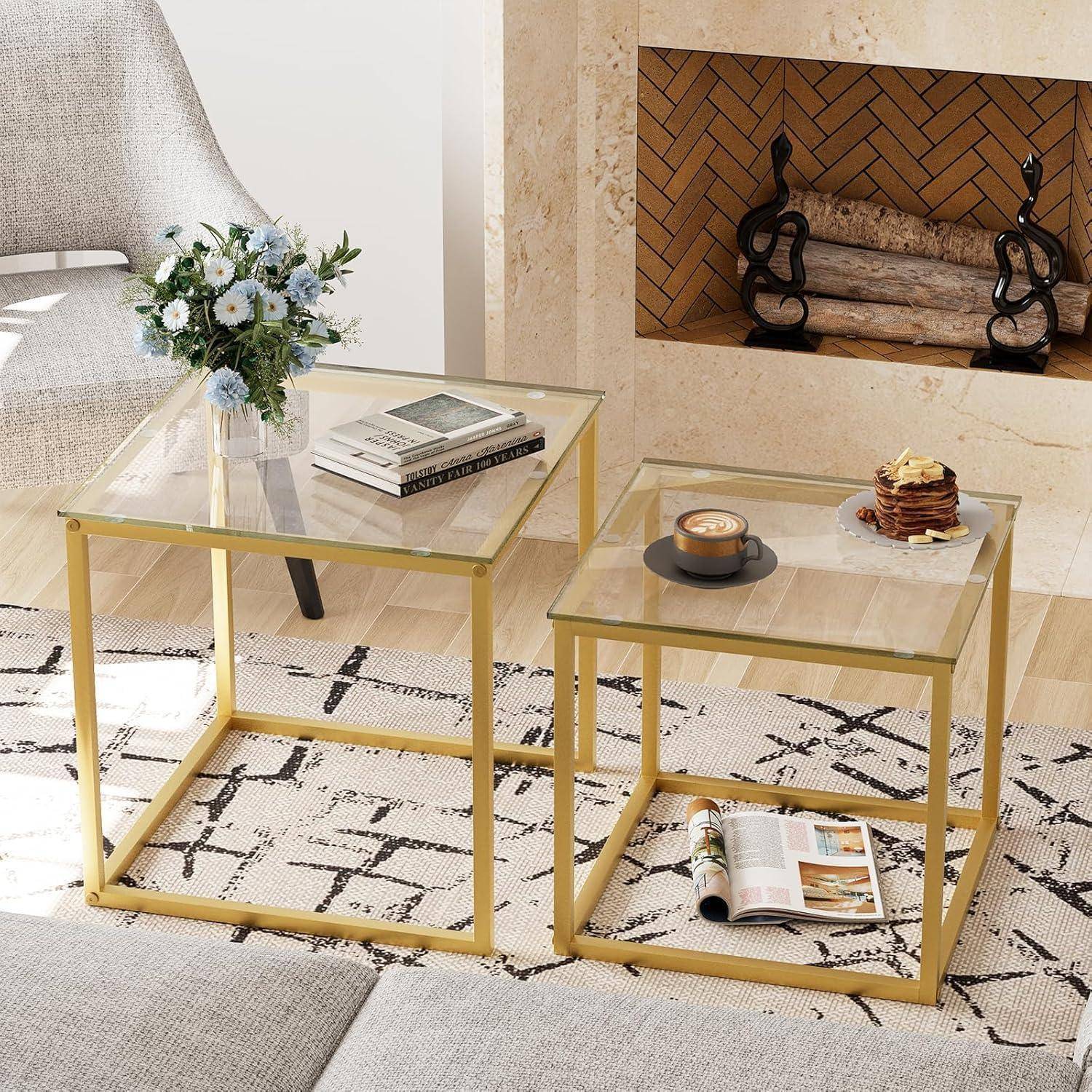 Zwei Beistelltische aus Glas und Gold auf einem gemusterten Teppich in der Nähe eines Kamins. Auf einem Tisch stehen Blumen und ein Buch, auf dem anderen eine Kerze und ein Dessert.