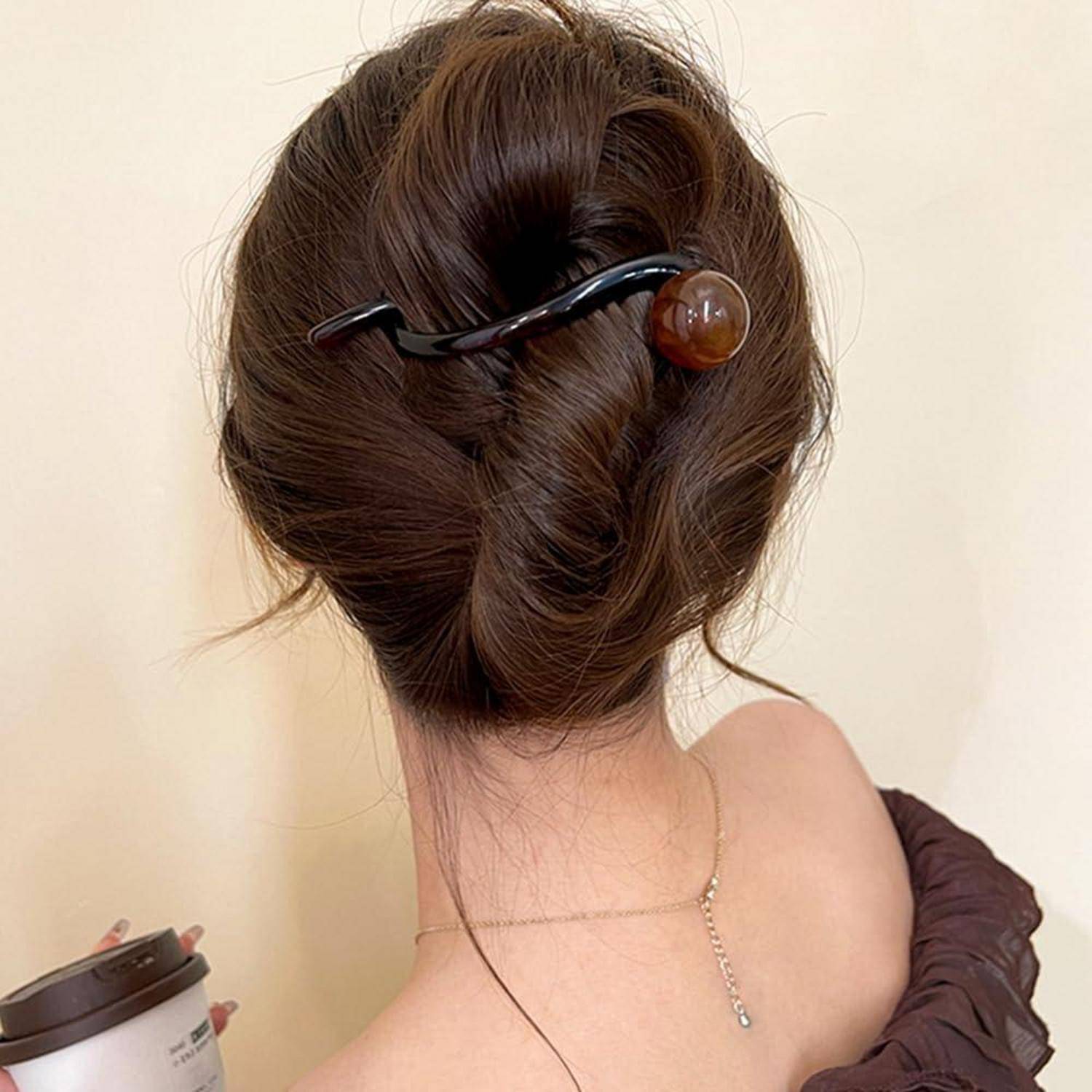 Eine Frisur mit einem ordentlichen Dutt, befestigt mit einer schwarzen Haarspange mit einer braunen Perle; eine Hand, die im Hintergrund eine Kaffeetasse hält.