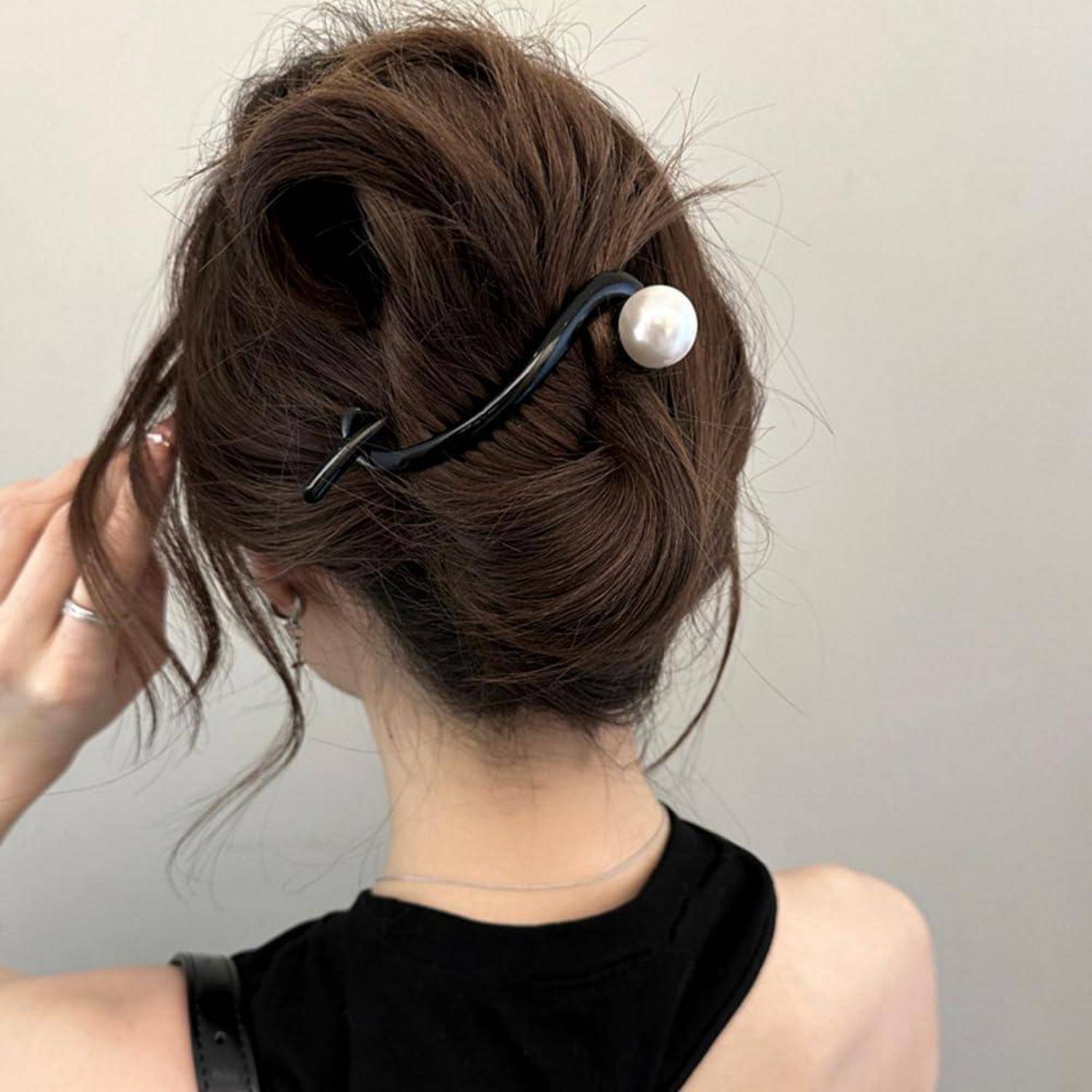Eine Person mit braunen Haaren in einem Dutt, gesichert mit einer dekorativen Perlen-Haarspange. Sie trägt ein schwarzes, ärmelloses Top.