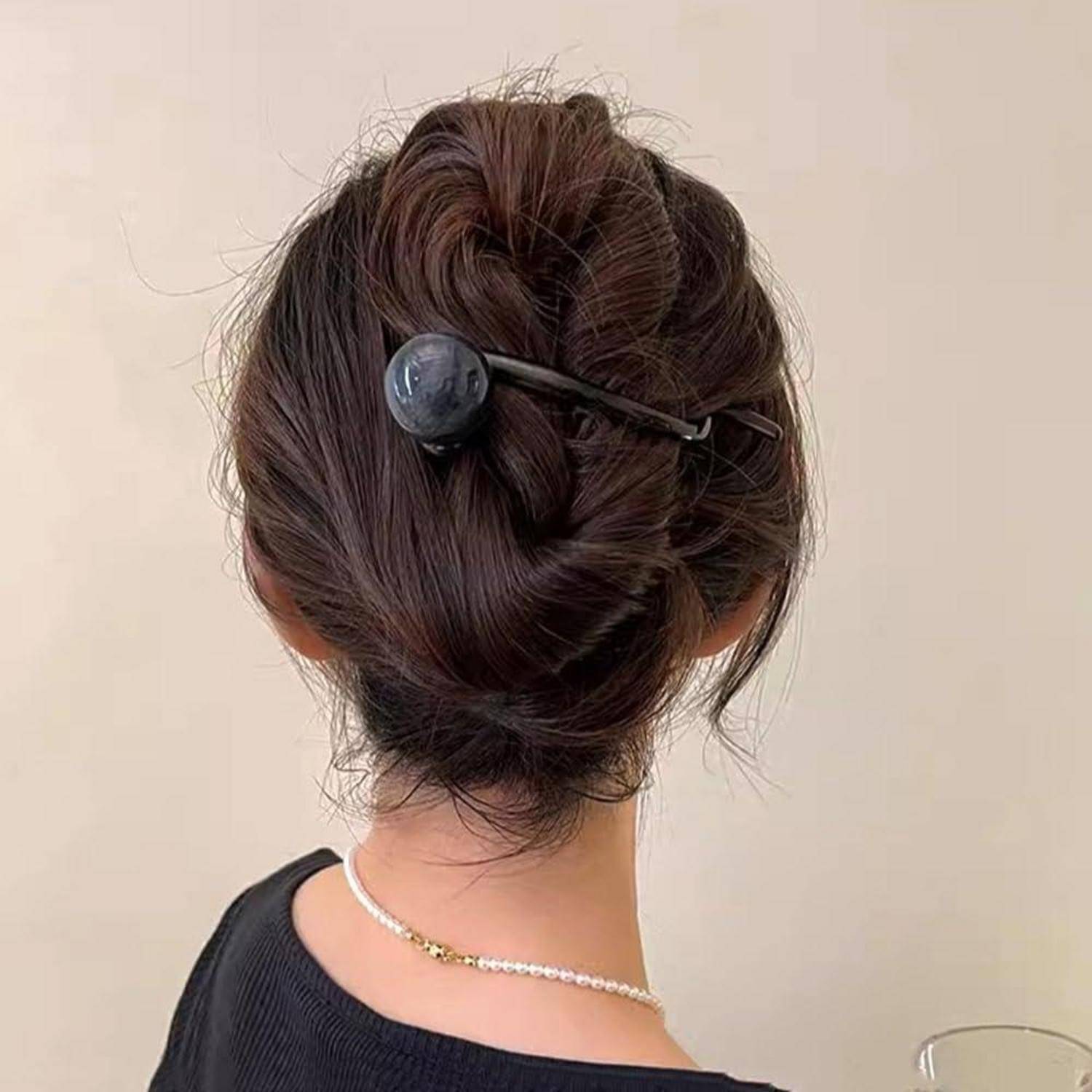 Eine Frau mit hochgestecktem Haar in einem eleganten Dutt, befestigt mit einer schwarzen Haarnadel, trägt ein schwarzes Oberteil und eine zarte Halskette.