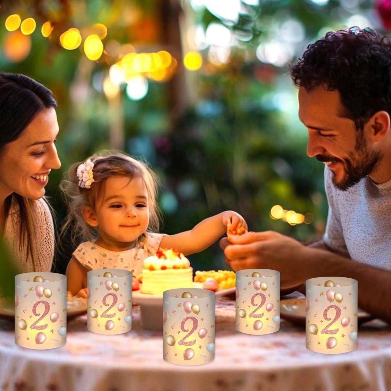 Eine Familie feiert den Geburtstag eines Kindes im Freien, mit einer Torte und brennenden Kerzen, die die Zahl '2' zeigen. Die Atmosphäre ist festlich und voller Freude.