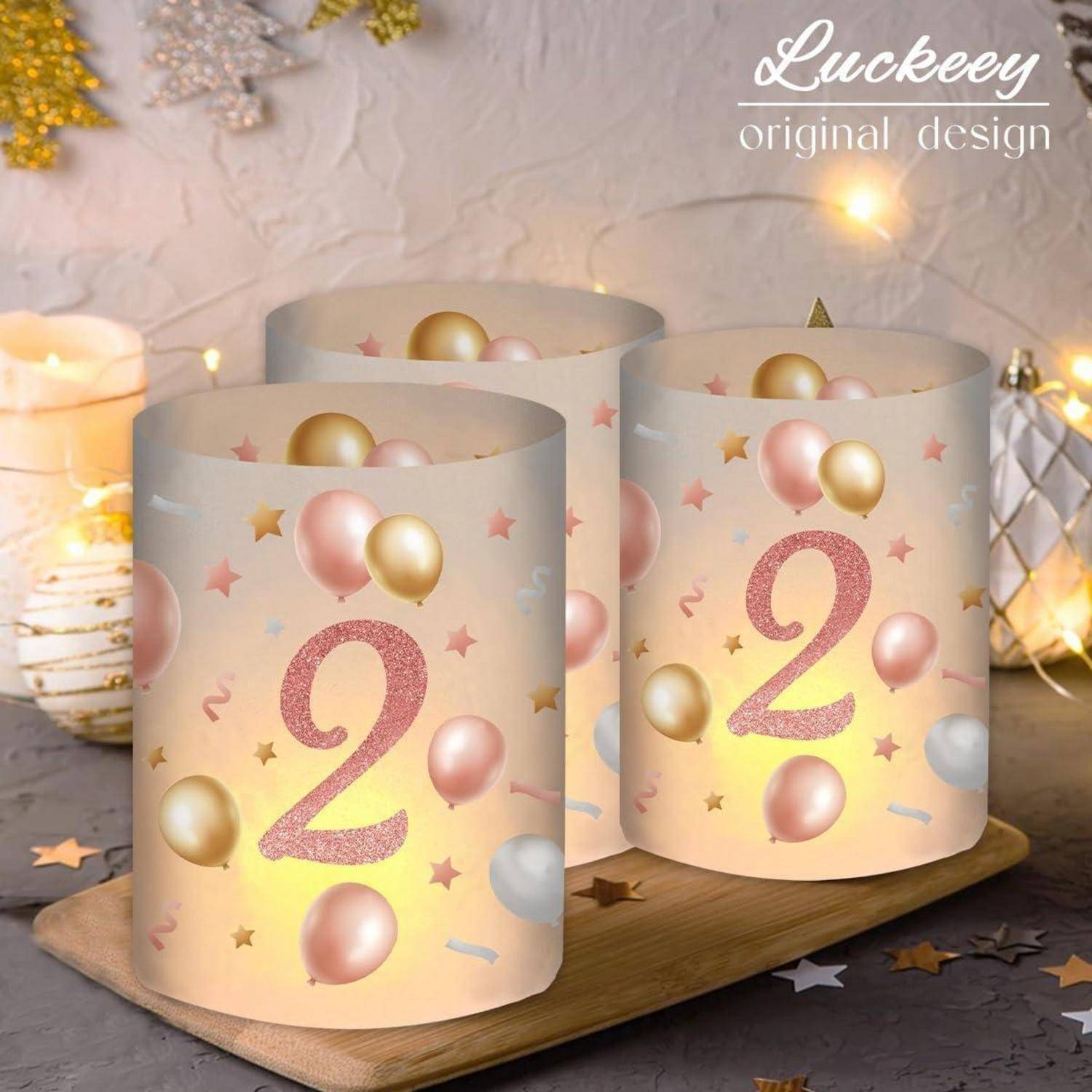 Drei dekorative Kerzenhalter mit rosa '2' und Ballondesigns auf einem Holztablett, vor einem rustikalen Hintergrund mit Lichterketten.