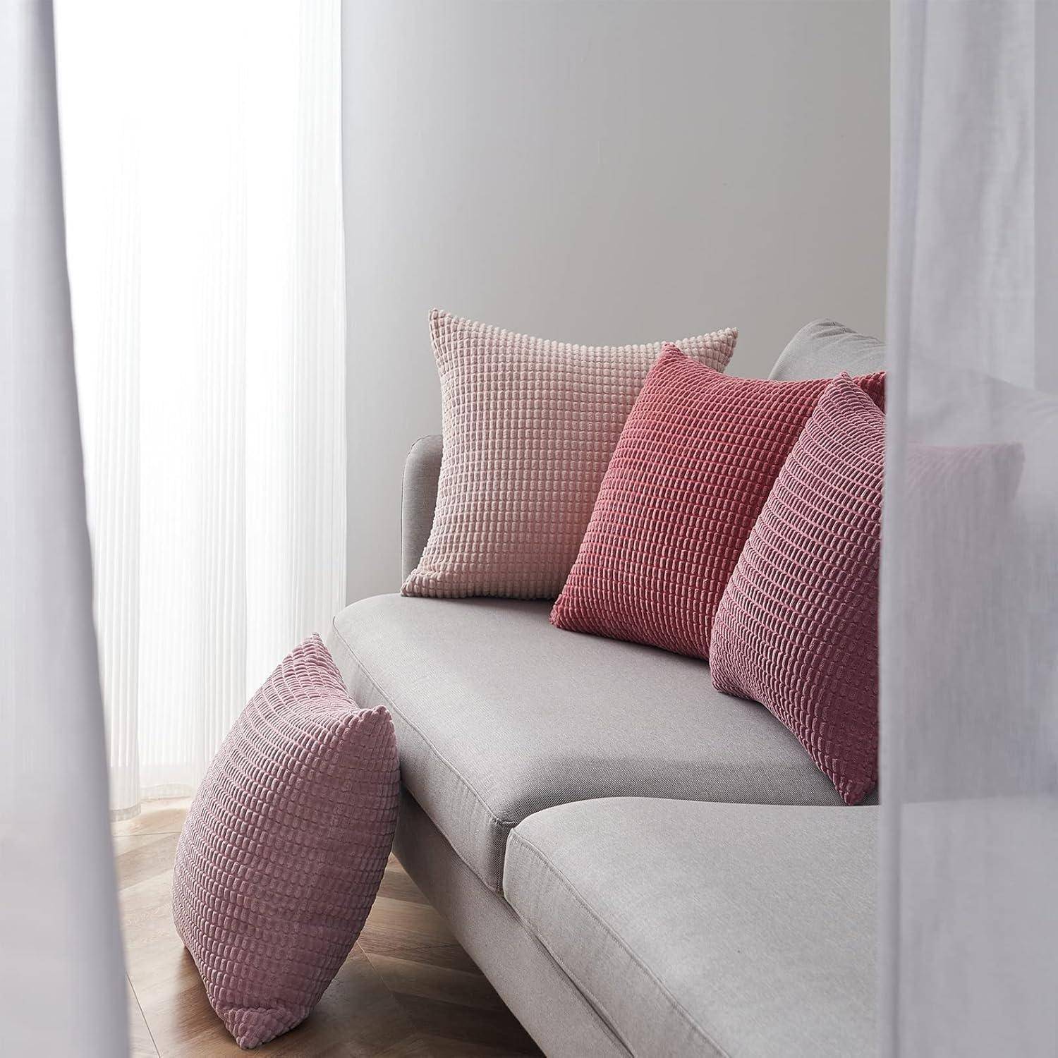 Eine gemütliche Ecke eines Wohnzimmers mit einem hellgrauen Sofa, auf dem drei rosafarbene Kissen mit Strukturmuster liegen, nahe einem Fenster mit zarten weißen Gardinen.