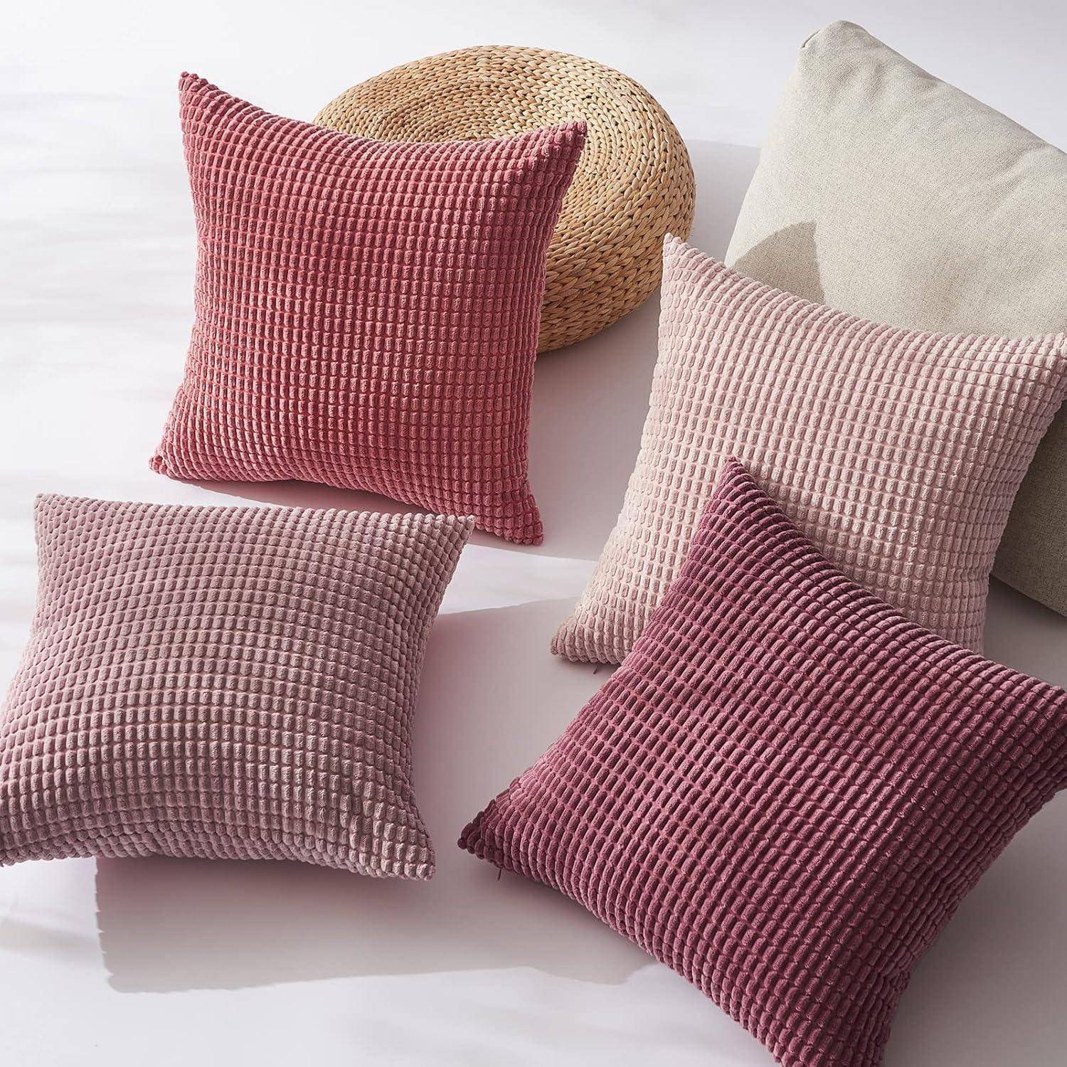 Vier rosa und beige strukturierte Kissen, die auf einer hellen Oberfläche angeordnet sind, mit einem Strohhut und einem beigen Kissen im Hintergrund, wodurch eine gemütliche Atmosphäre entsteht.