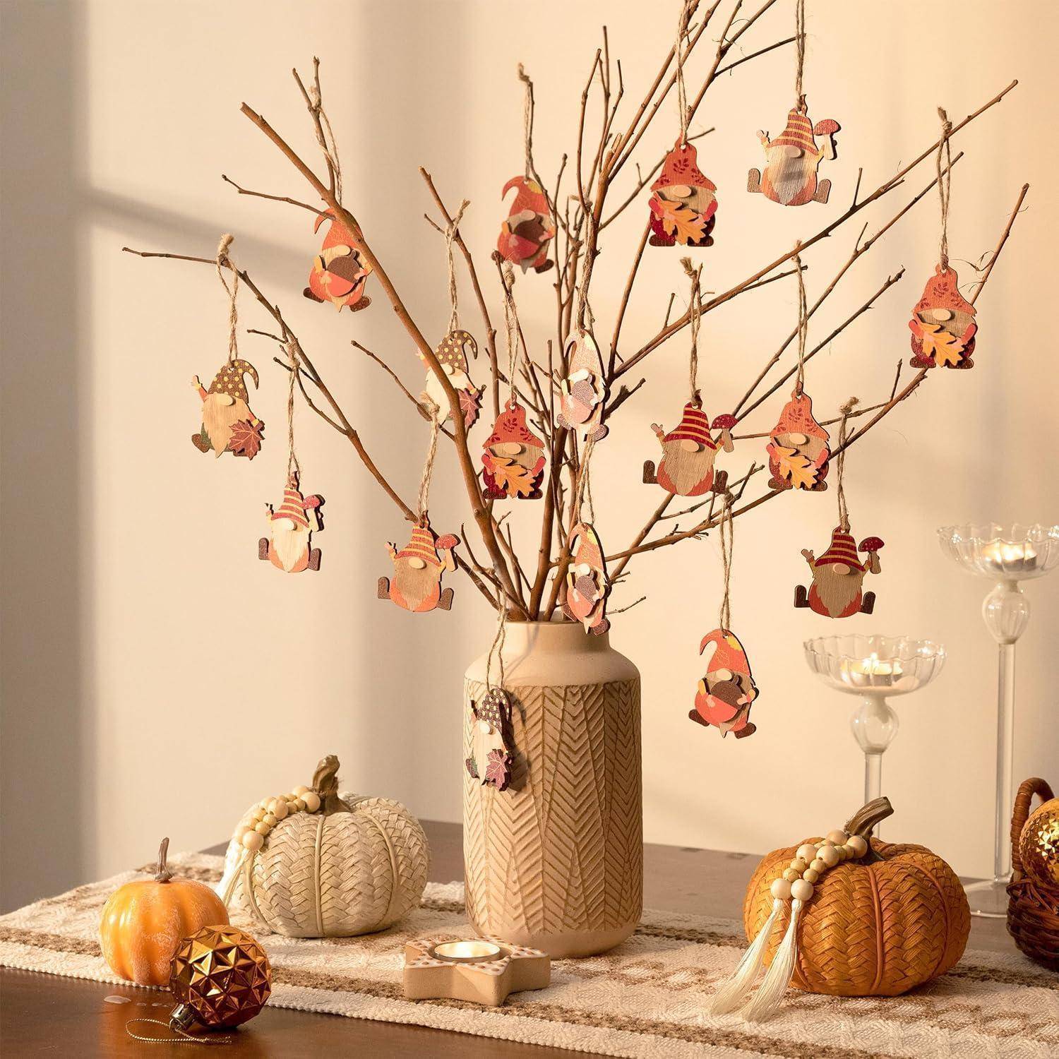 Eine Keramikvasevase enthält kahle Zweige, die mit herbstlichen Ornamenten geschmückt sind. In der Nähe befinden sich dekorative Kürbisse und Glaskerzenhalter auf einem Tisch.