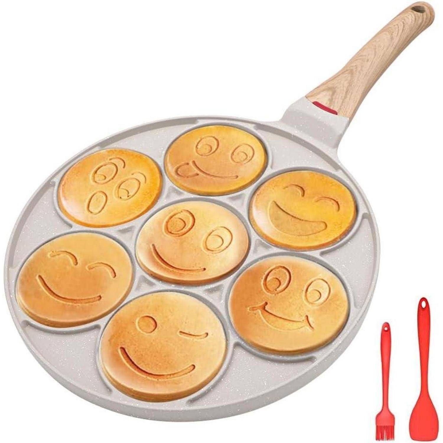 Eine Pfannkuchenpfanne mit sechs Smiley-Gesichts-Formen und zwei roten Küchenutensilien daneben.