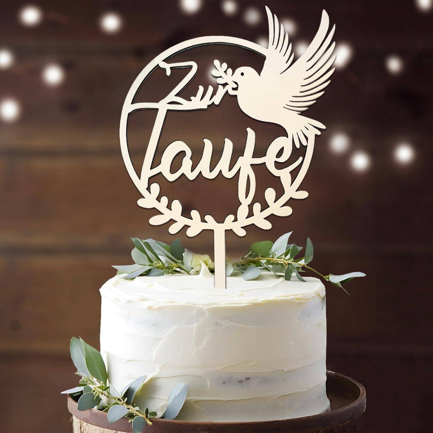 Eine weiße Torte mit grünen Blättern oben, verziert mit einem dekorativen Topper, der eine Taube und das Wort 'Taufe' zeigt, vor einem Holzhintergrund.