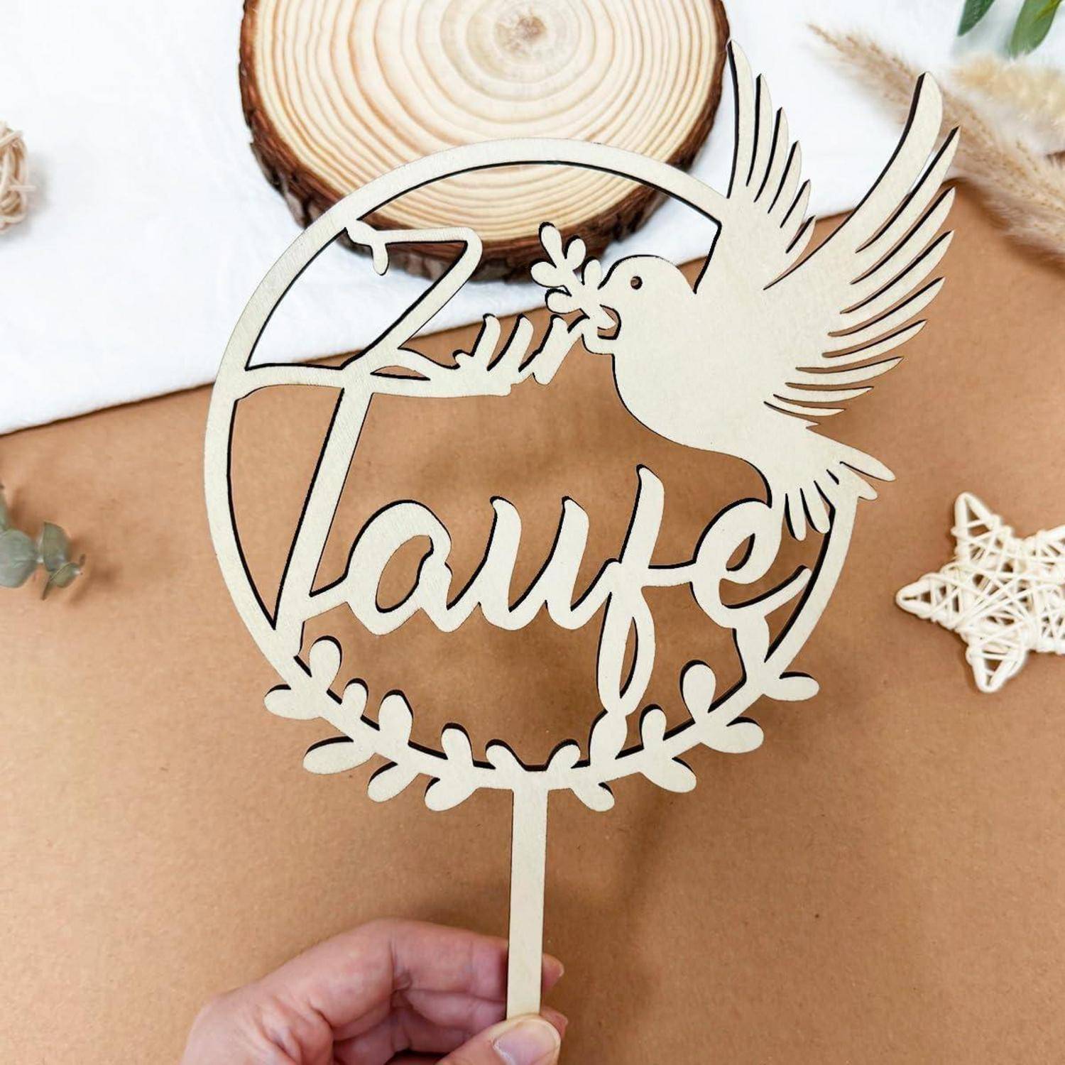 Ein Holz-Cake Topper mit einer Taube, die einen Olivenzweig trägt, und dem Wort 'Taufe', gehalten von einer Hand.