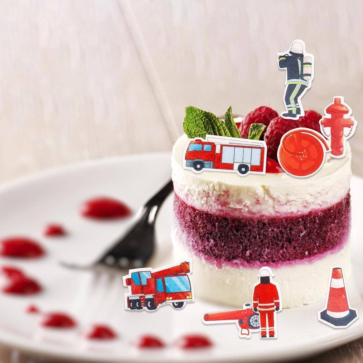 Ein Stück roter Samtkuchen mit weißer Buttercreme ist mit Papierschnipseln eines Feuerwehrautos, eines Feuerwehrmanns und eines Hydranten auf einem weißen Teller geschmückt.