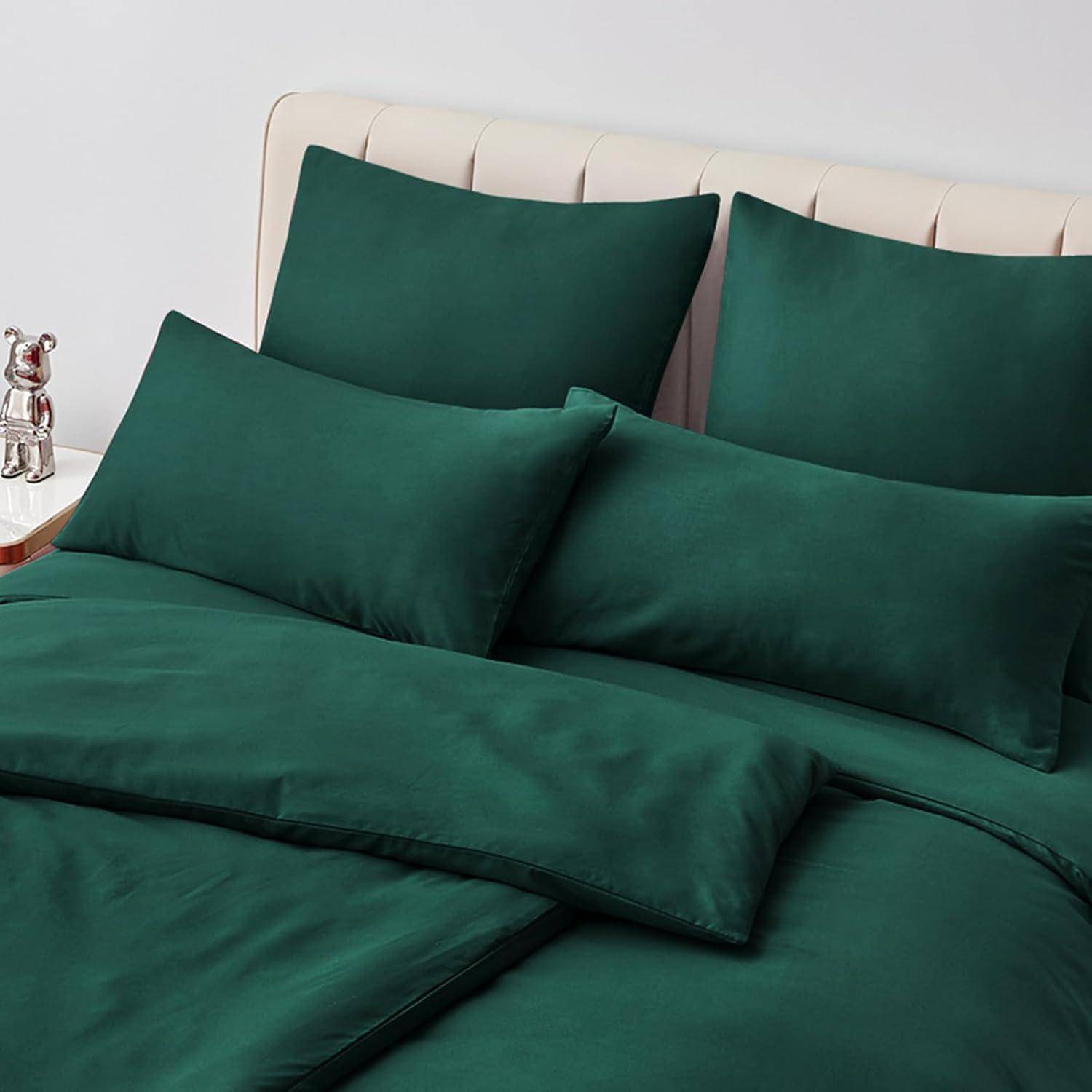 Ein ordentlich gemachtes Bett mit dunkelgrünen Laken, Kissen und Bettdecke, vor einem hellbeigen gepolsterten Kopfteil. Eine kleine silberne Bärenfigur steht auf dem Nachttisch.