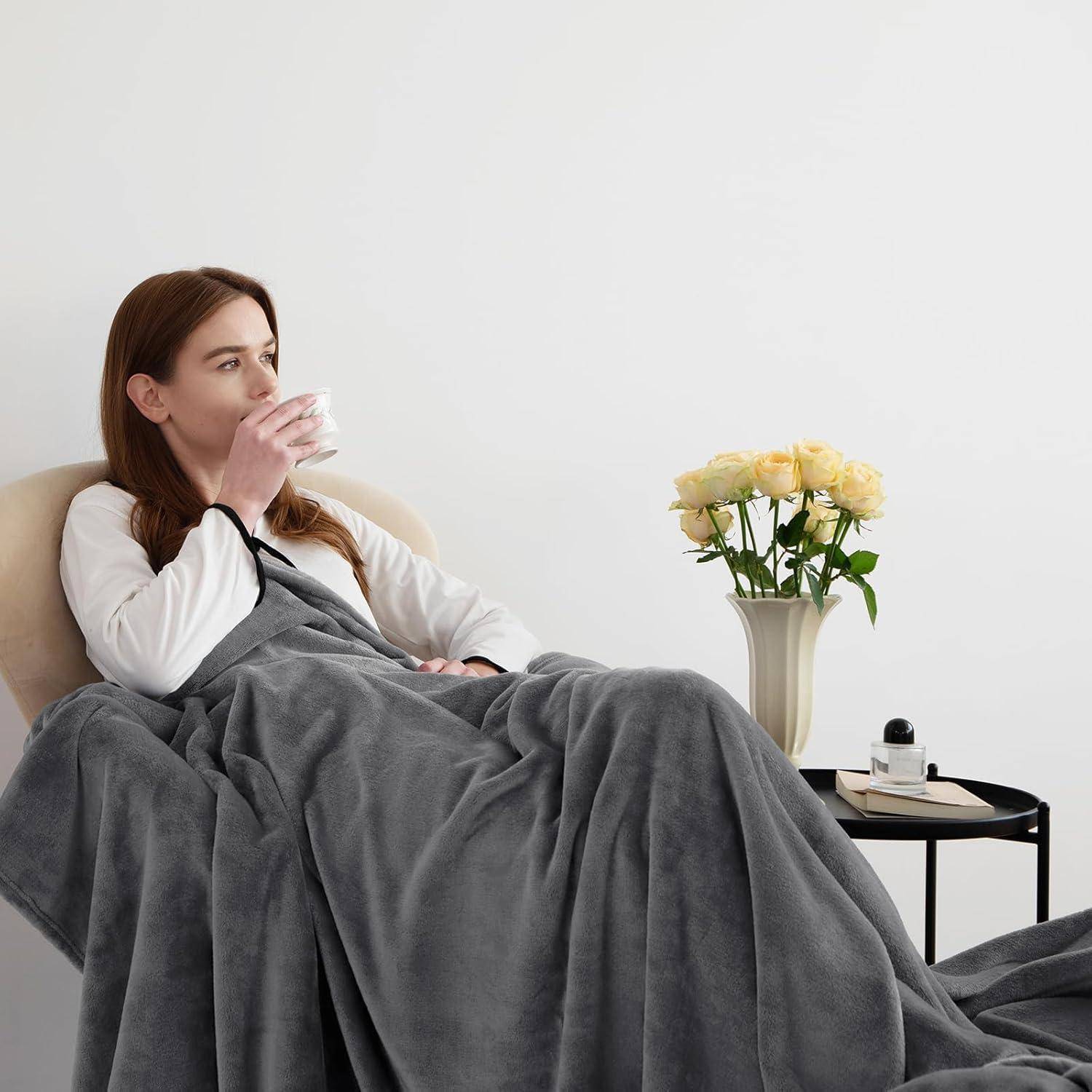 Eine Frau sitzt auf einem Sofa, in eine graue Decke gehüllt, und nippt an einem heißen Getränk aus einer Tasse. Auf dem Tisch daneben stehen eine Vase mit gelben Rosen und ein Buch.