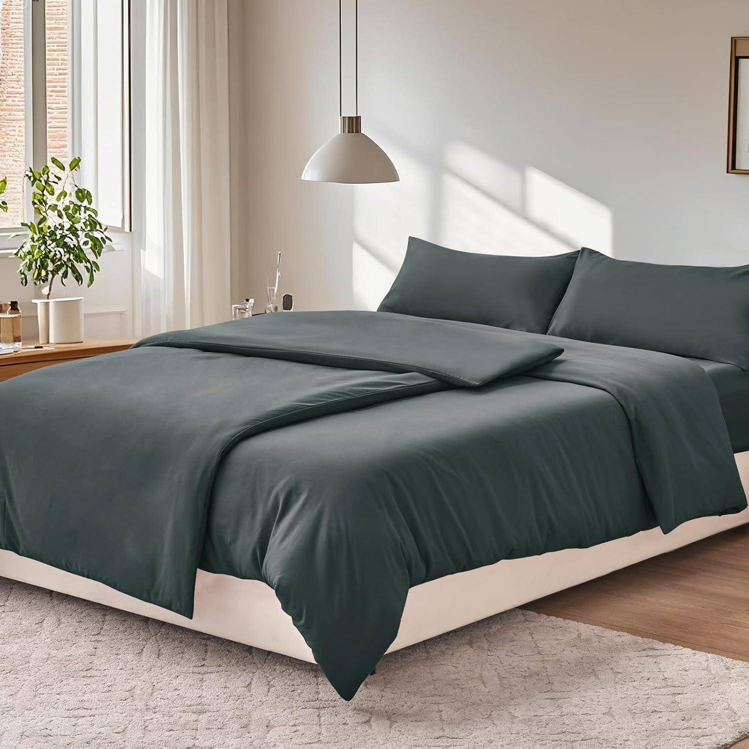 Ein ordentlich gemachtes Bett mit dunkelgrauer Bettwäsche, zwei Kissen und einer Bettdecke. Ein Holznachttisch mit einer Pflanze und Büchern in einem hell erleuchteten Schlafzimmer.