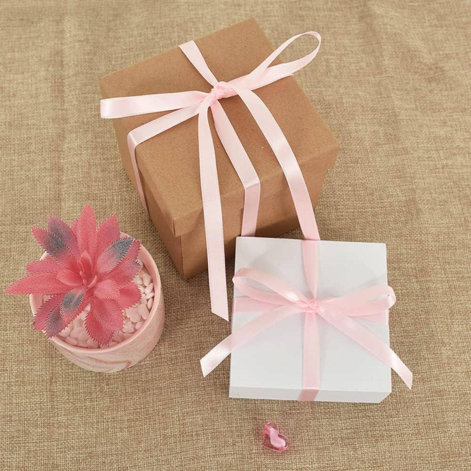 Eine braune Geschenkbox und eine weiße Geschenkbox, beide mit rosa Bändern gebunden, stehen auf einer Juteoberfläche neben einer dekorativen Blume in einem rosa Topf.