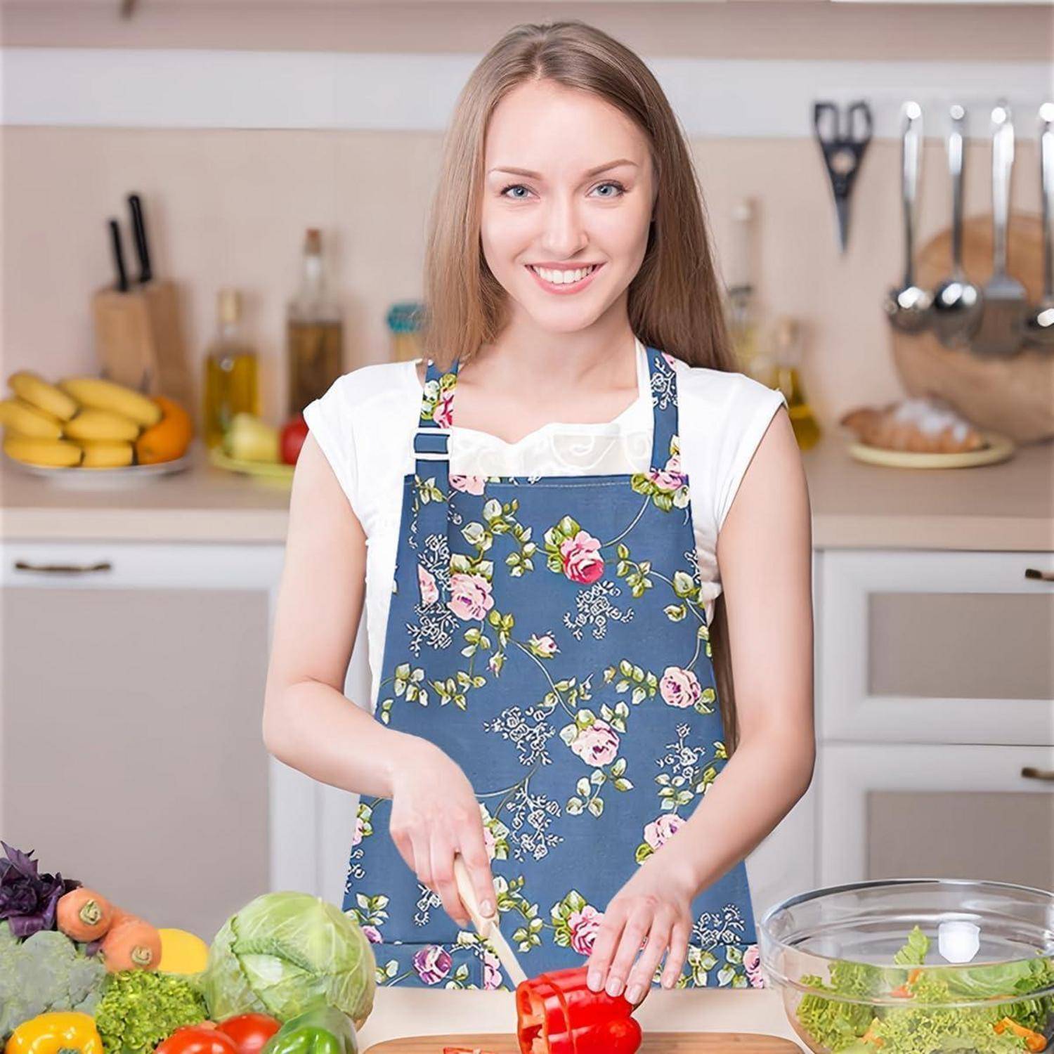 Eine Frau in einer Blumenschürze lächelt, während sie eine rote Paprika in einer Küche mit verschiedenen Gemüsesorten und Kochutensilien um sie herum schneidet.
