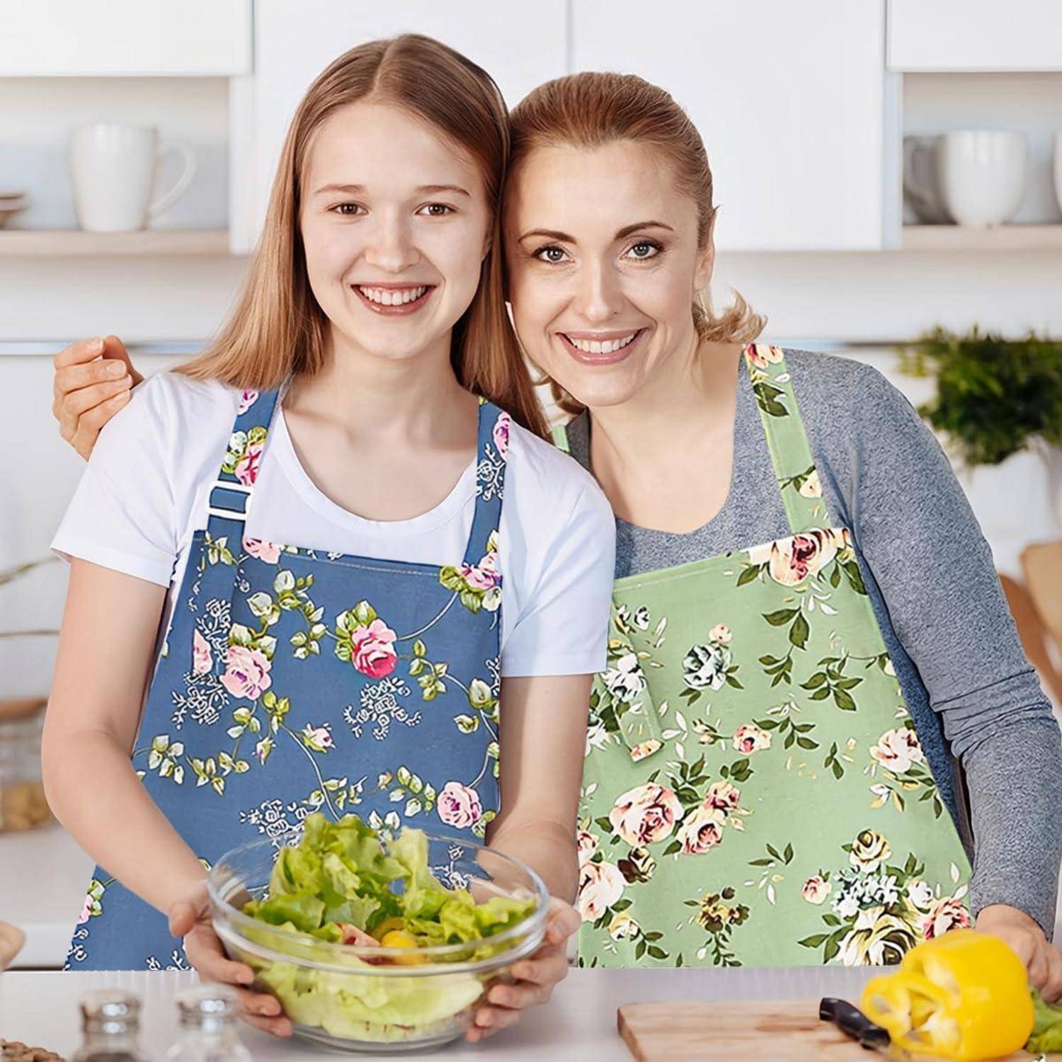 Eine Mutter und ihre Tochter, beide mit Blumenschürzen bekleidet, lächeln, während sie in einer Küchenumgebung einen Salat zubereiten.