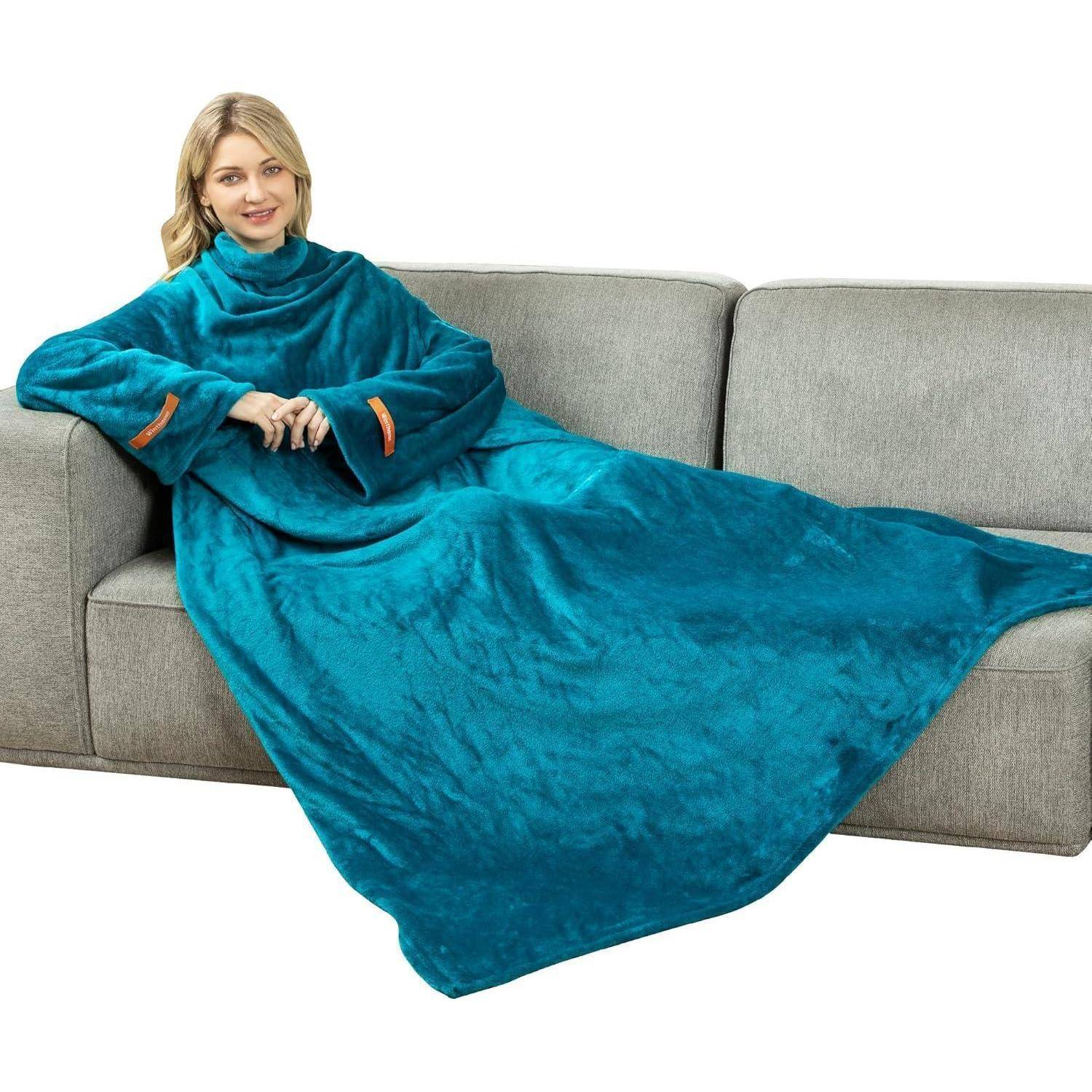 Eine Person sitzt auf einem grauen Sofa und trägt einen türkisfarbenen Wearable-Blanket mit Ärmeln, sieht behaglich und entspannt aus.