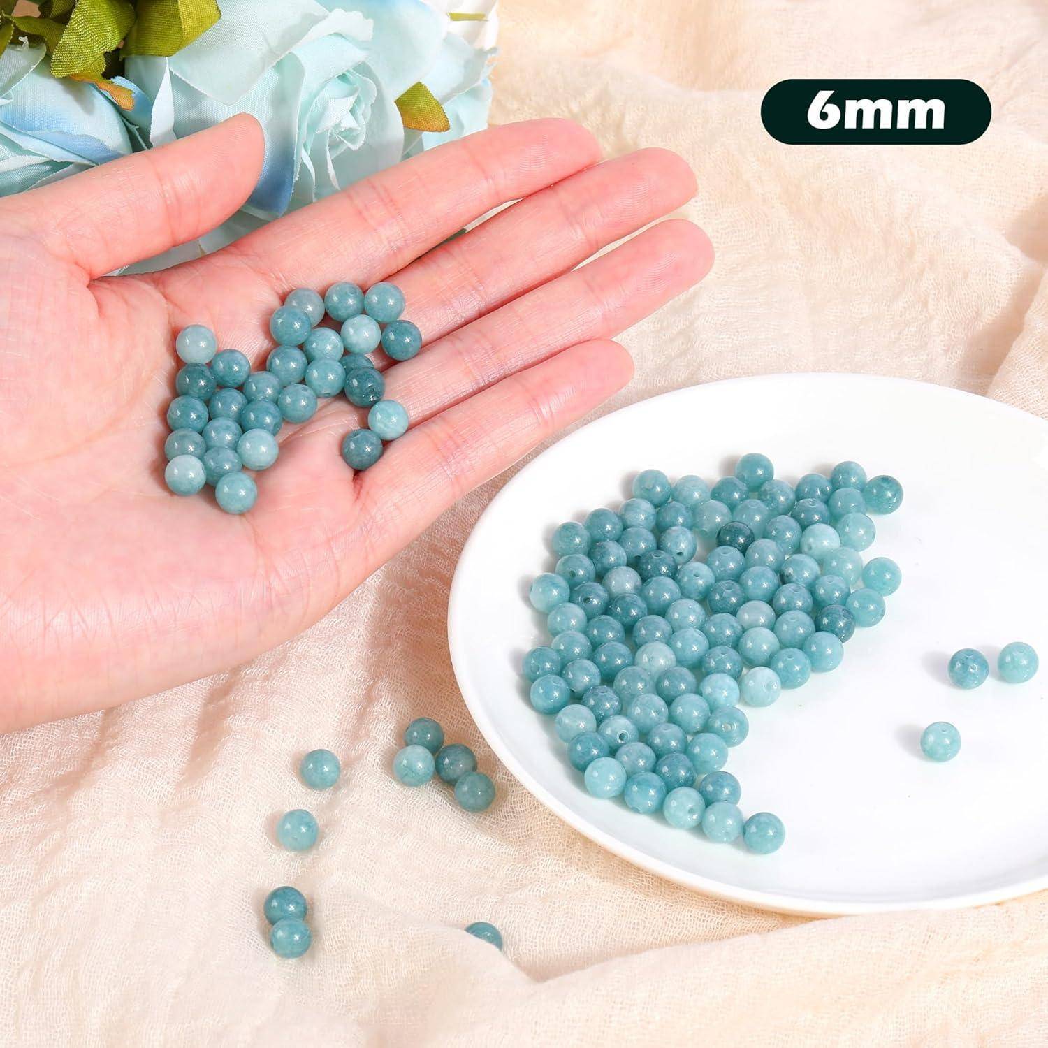 Eine Hand, die mehrere 6-mm-azurblaue Perlen hält, neben einem weißen Teller, der weitere Perlen der gleichen Art enthält. Blaue Blumen sind teilweise sichtbar.