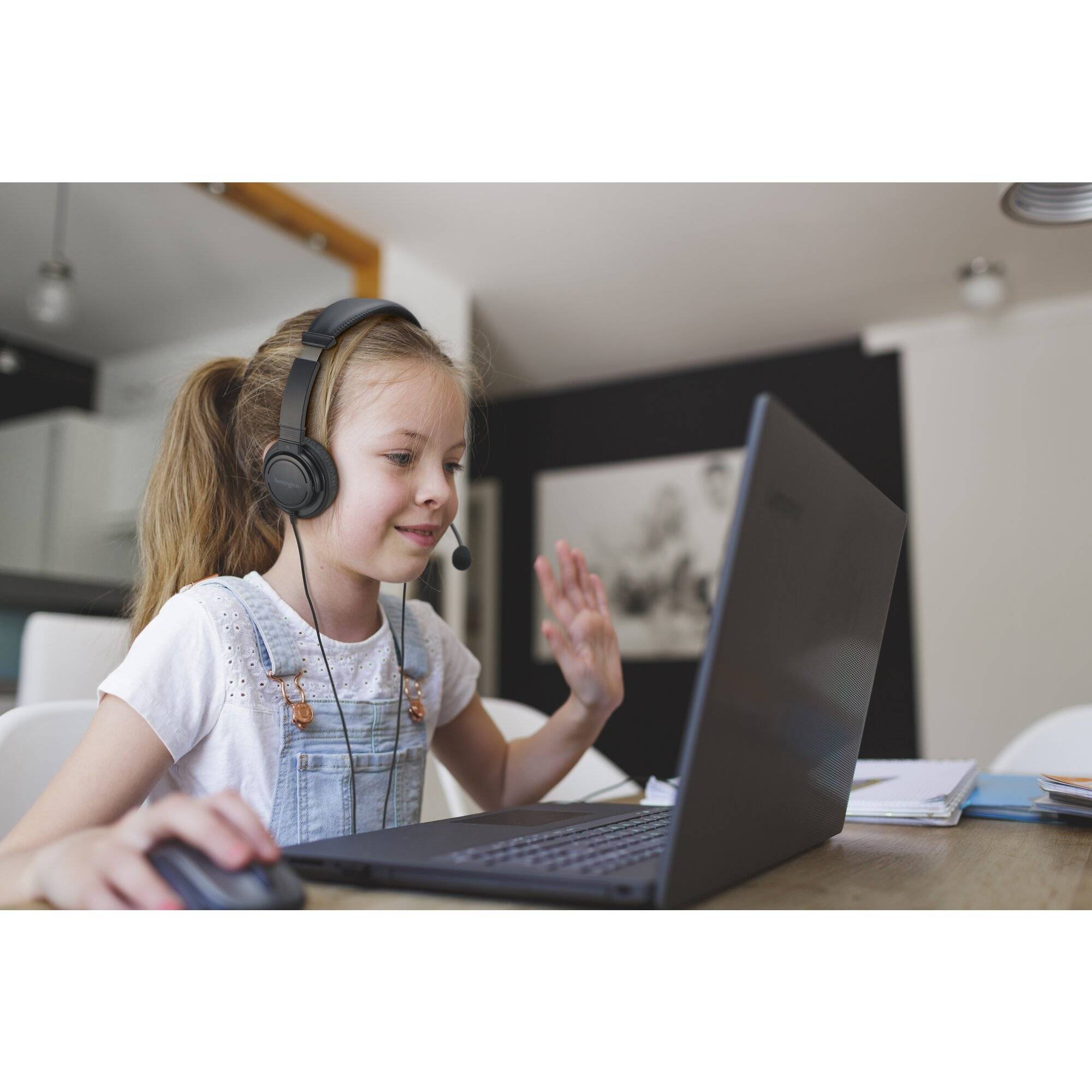 Junges Mädchen mit Kopfhörer winkt, während es einen Laptop auf einem Schreibtisch während eines Online-Anrufs oder einer Online-Lektion nutzt.