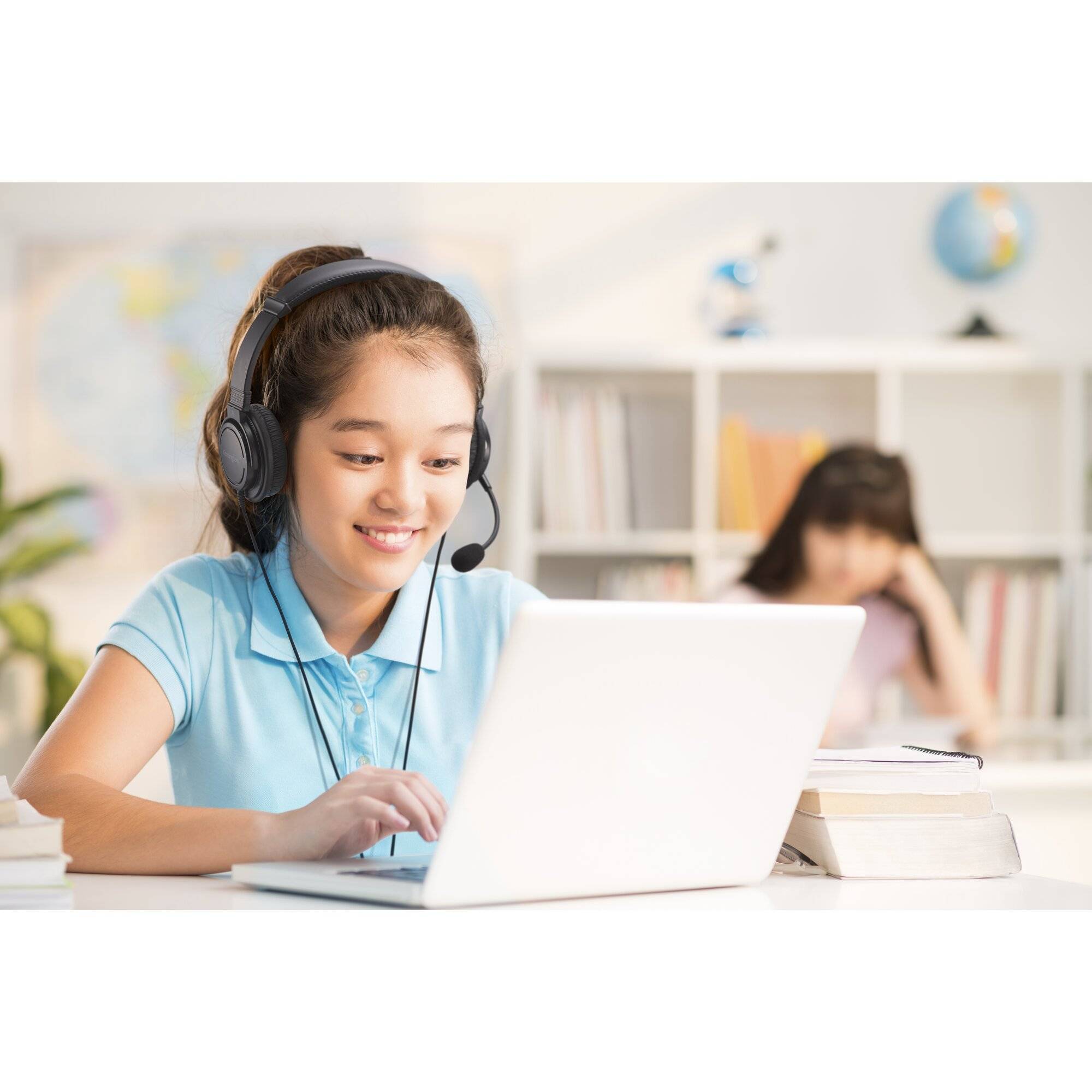 Mädchen mit Kopfhörer nutzt einen Laptop an einem Schreibtisch, mit Büchern in der Nähe und einem unscharfen Studenten, der im Hintergrund arbeitet.