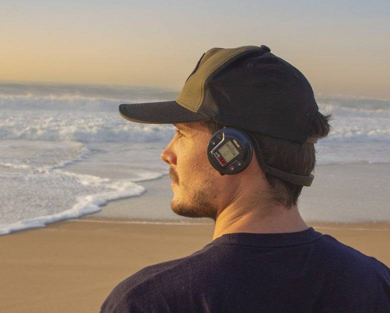Eine Person mit Kopfhörern und Mütze blickt während des Sonnenuntergangs auf einen sandigen Strand auf das Meer.