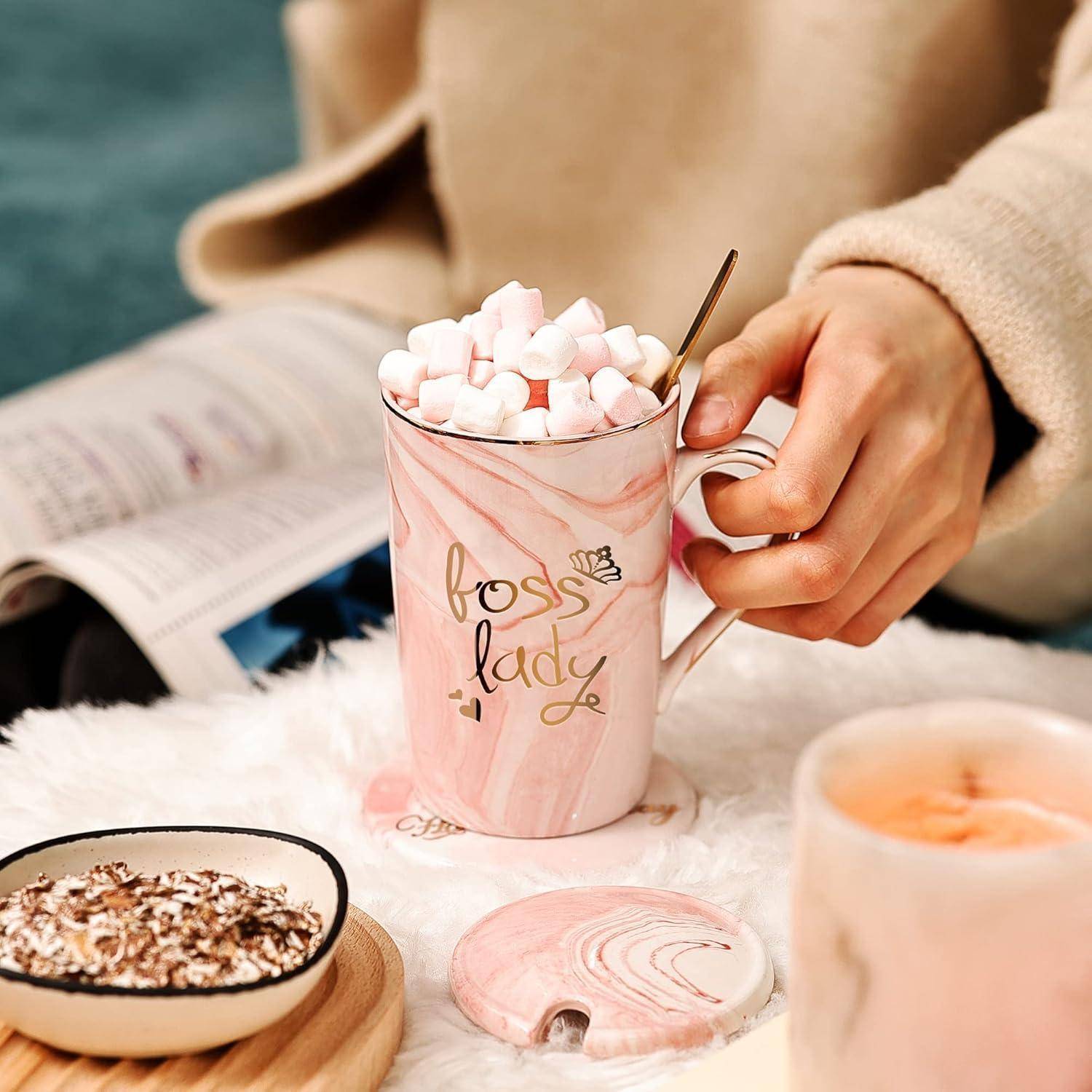 Eine Person, die eine rosa Tasse mit dem Aufdruck 'Boss Lady' hält, gefüllt mit Marshmallows. Eine gemütliche Umgebung mit einer Zeitschrift und Snacks in der Nähe.