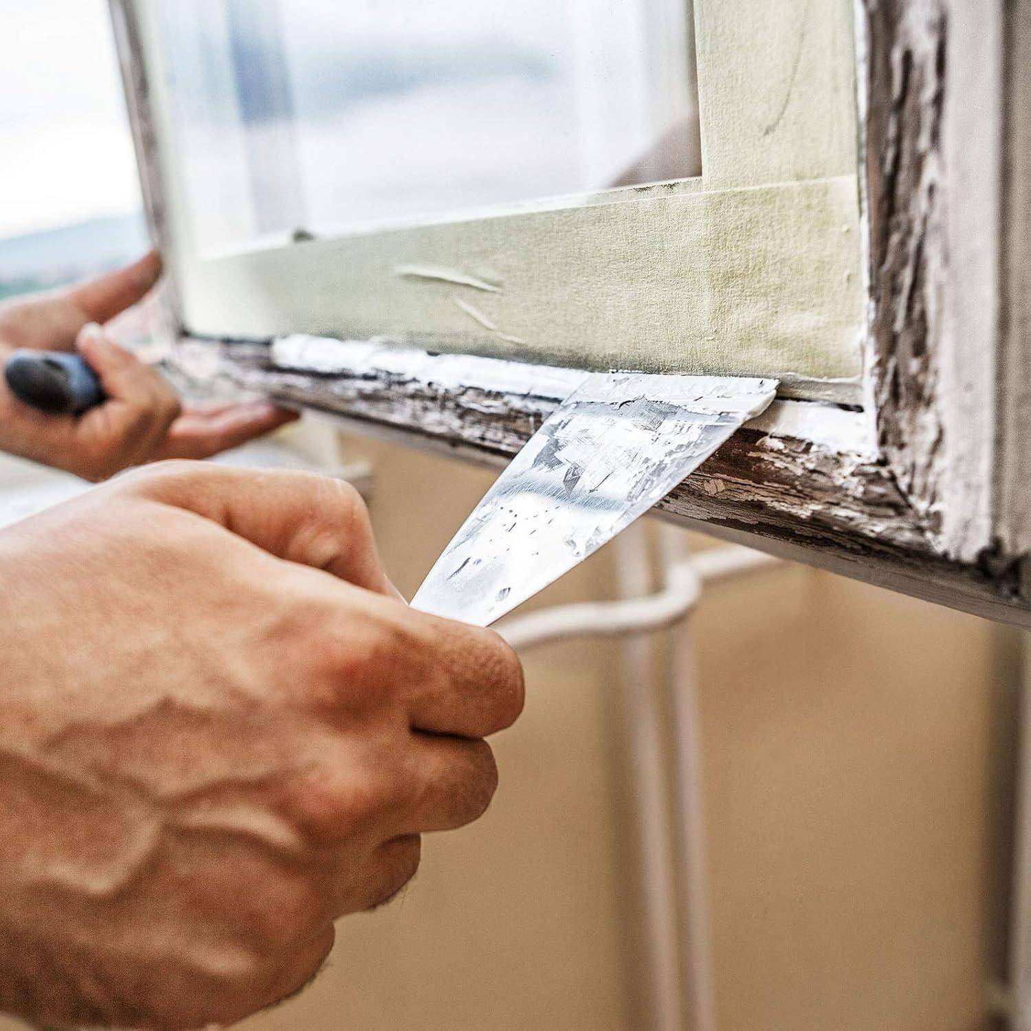 Eine Person, die alte Farbe von einem Fensterrahmen mit einem Spachtel abkratzt und die Oberfläche für das erneute Anstreichen vorbereitet.