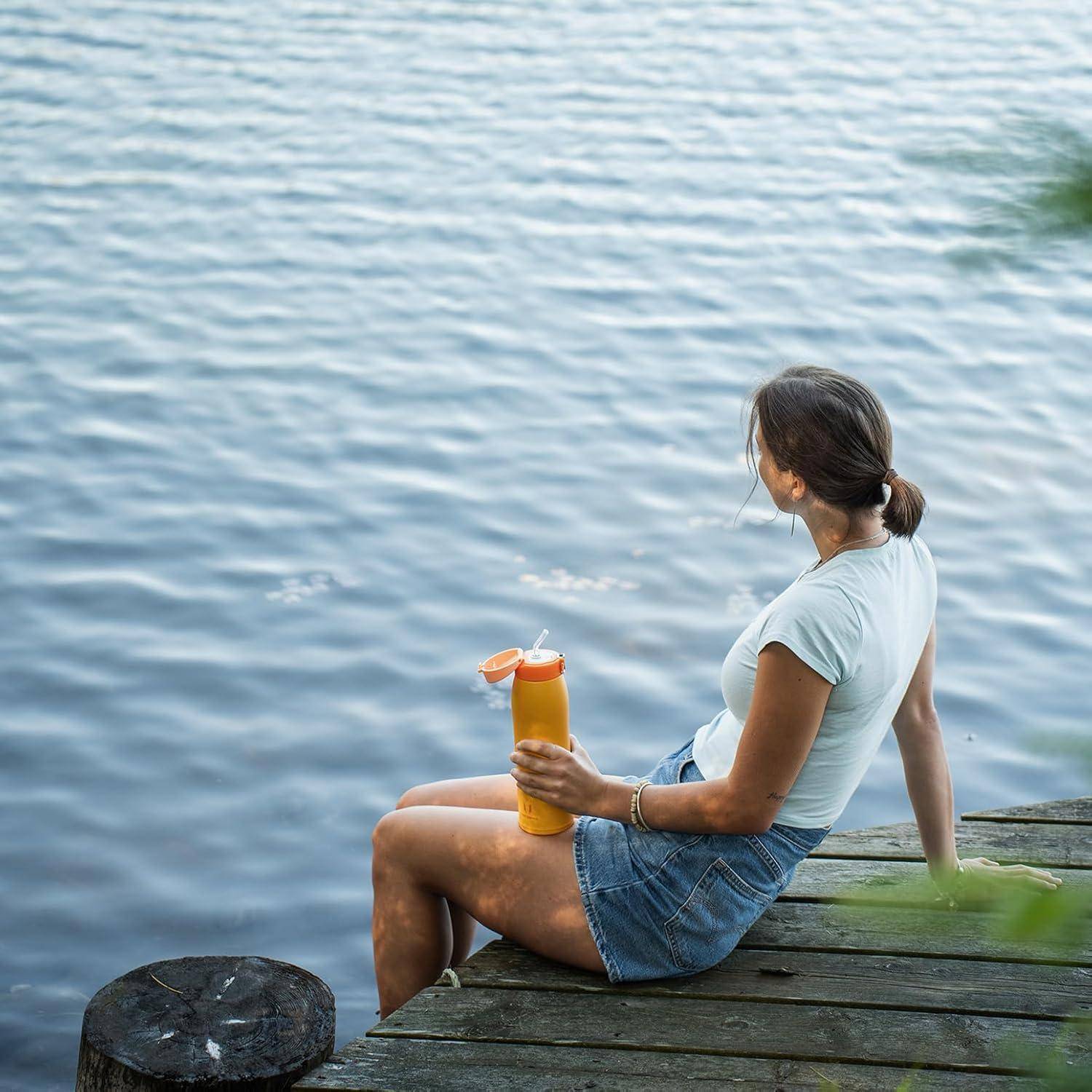Eine Person sitzt auf einem Holzsteg an einem ruhigen See und hält eine Flasche mit einem orangefarbenen Deckel, trägt lässige Sommerkleidung.