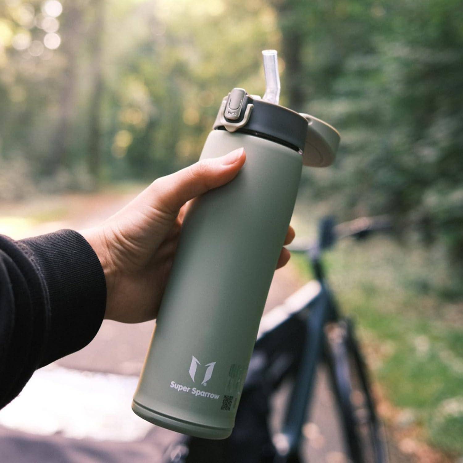 Eine Hand, die eine grüne Wasserflasche mit Strohdeckel hält, die mit 'Super Sparrow' markiert ist, vor einem unscharfen Hintergrund im Freien.