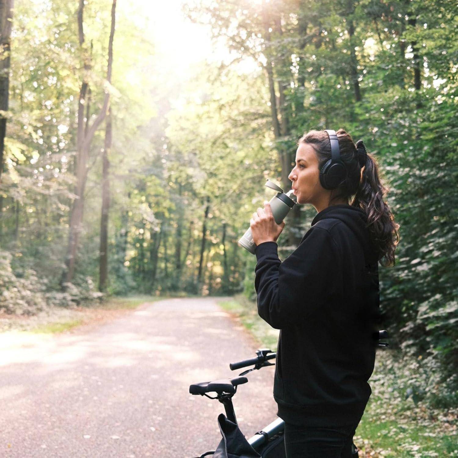 Eine Person mit Kopfhörern trinkt aus einer Flasche, während sie neben einem Fahrrad auf einem Waldweg steht, umgeben von hohen Bäumen bei Tageslicht.