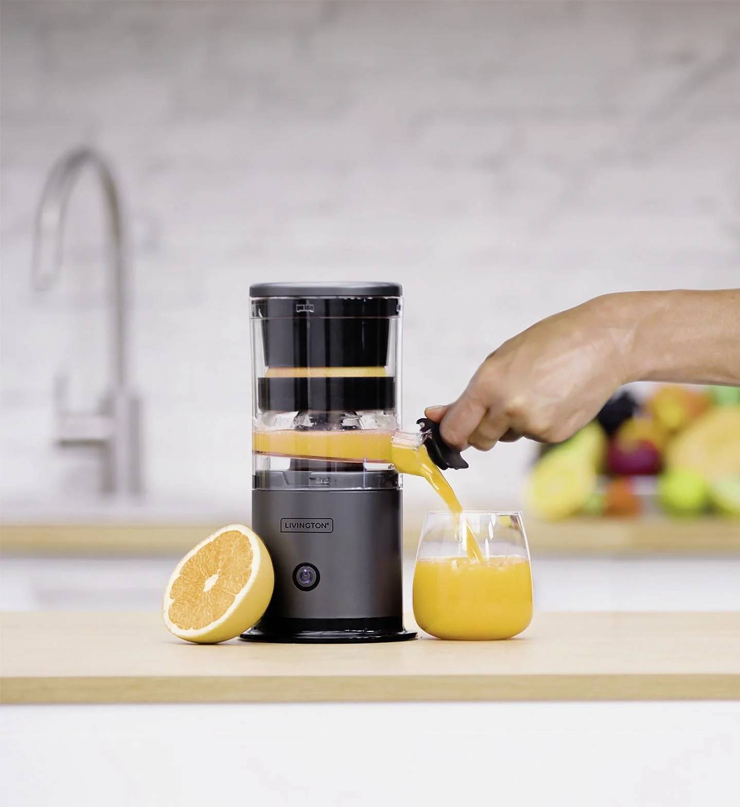 Eine Hand bedient einen Entsafter und presst Saft aus einer Orange in ein Glas auf einer Küchenarbeitsplatte. Im Hintergrund liegen eine halbierte Orange und Früchte.