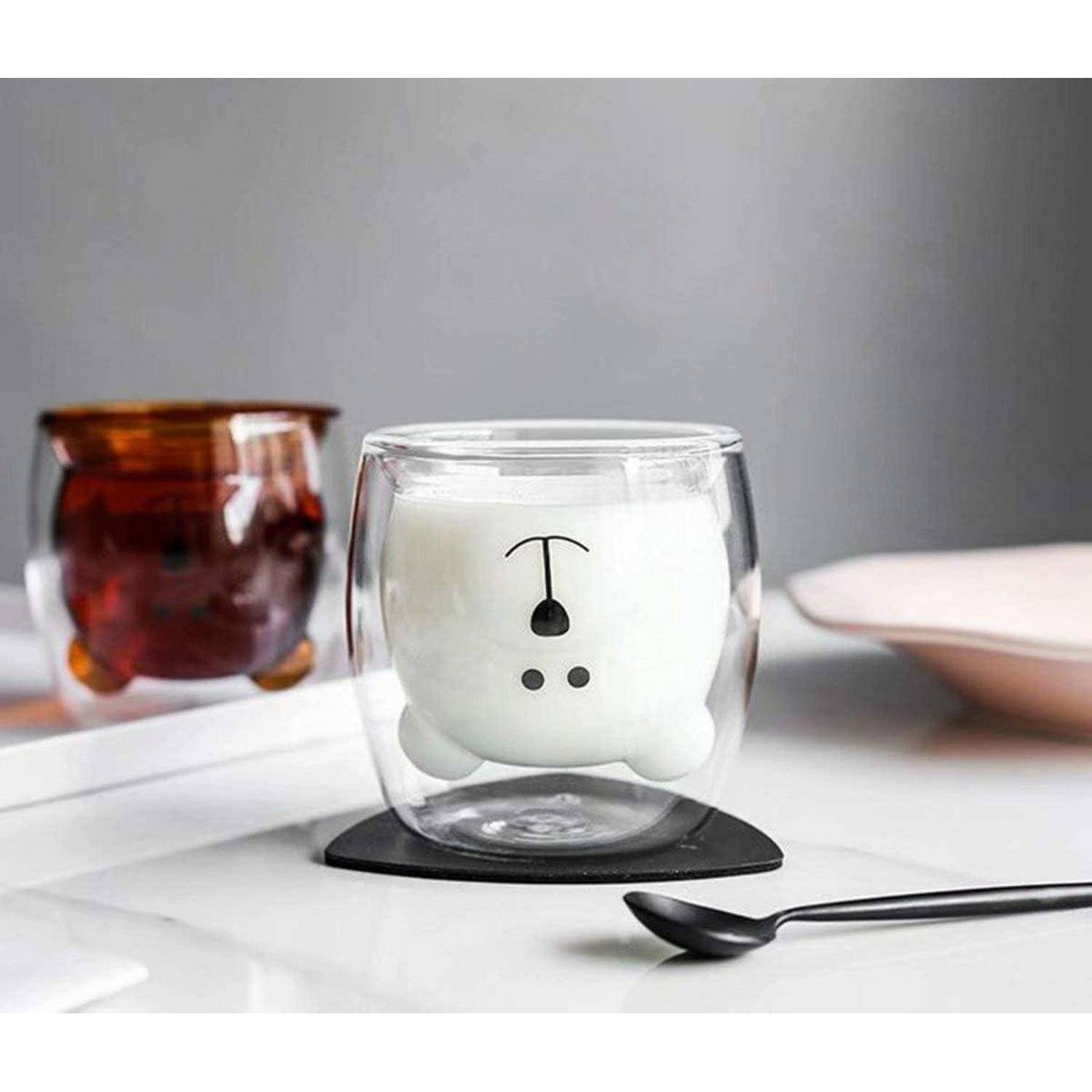 Eine doppelwandige Glasstasse in Bärengesichtsform, gefüllt mit Milch, mit einem weiteren Glas Tee im Hintergrund auf einer Arbeitsplatte.