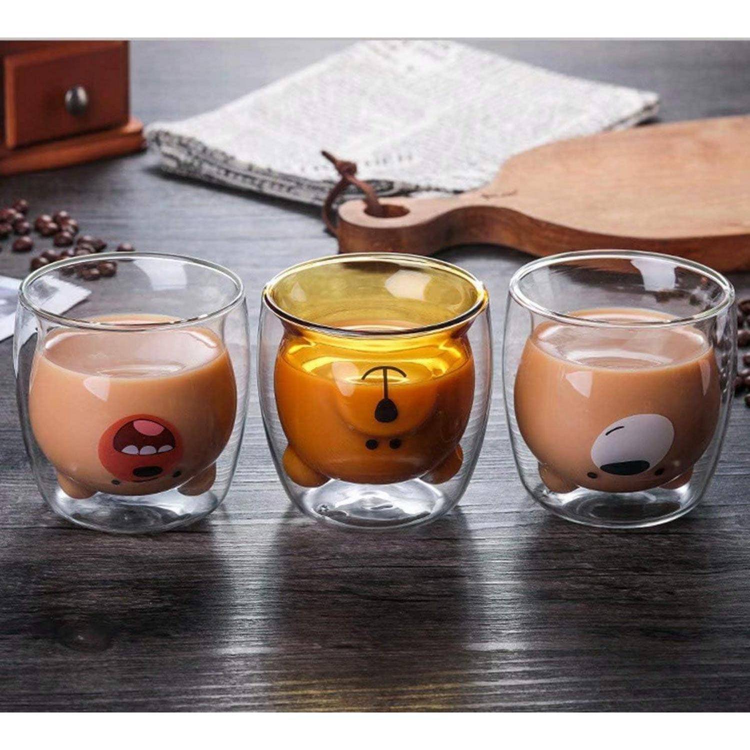 Drei durchsichtige Glastassen mit Comic-Tierfiguren enthalten eine beige Flüssigkeit. Die mittlere Tasse ist auf den Kopf gestellt und trägt ein orangefarbenes Bärendesign.