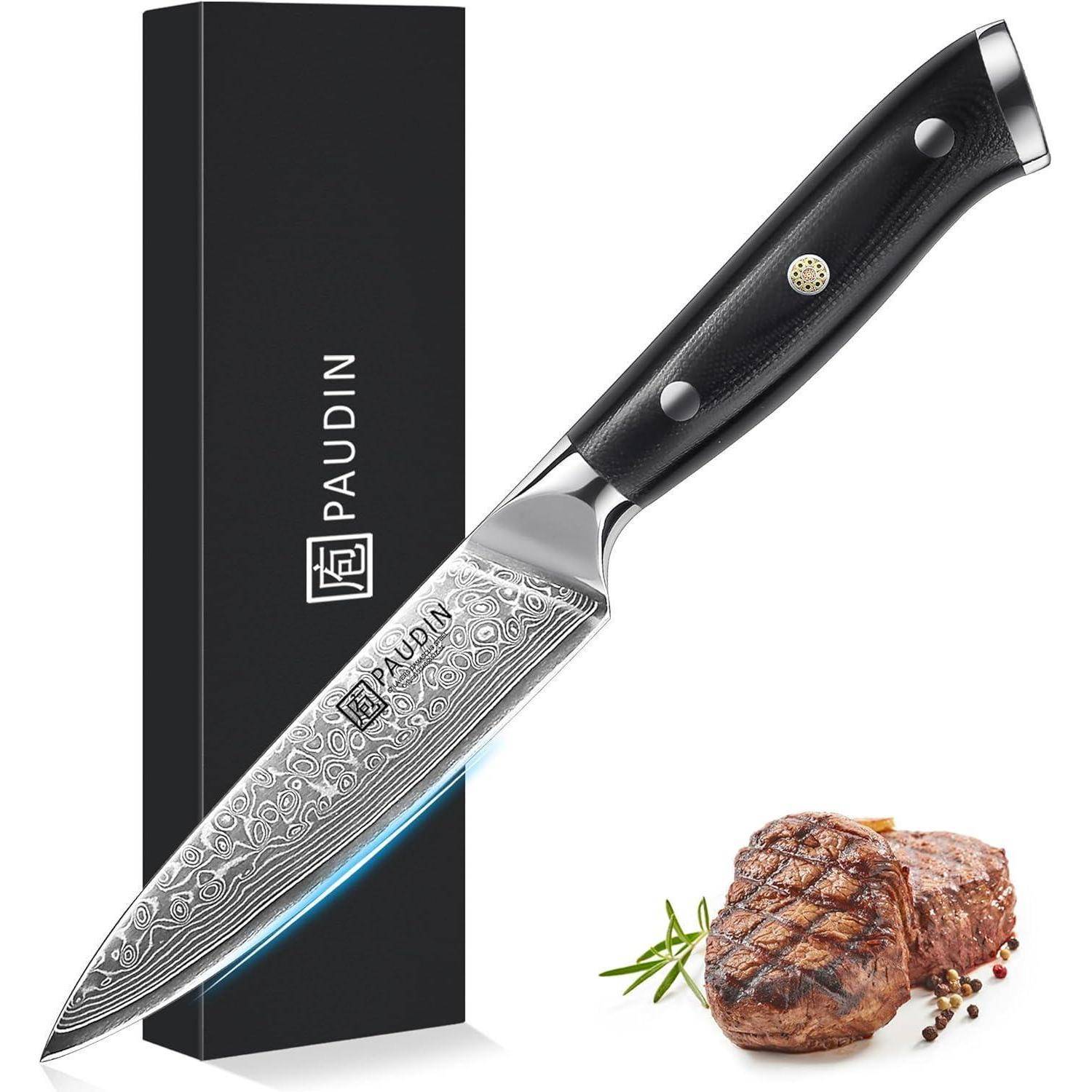 Ein Kochmesser mit gemusterter Klinge und schwarzem Griff neben einem gebratenen Steak, garniert mit Rosmarin, vor einer Schachtel mit der Aufschrift 