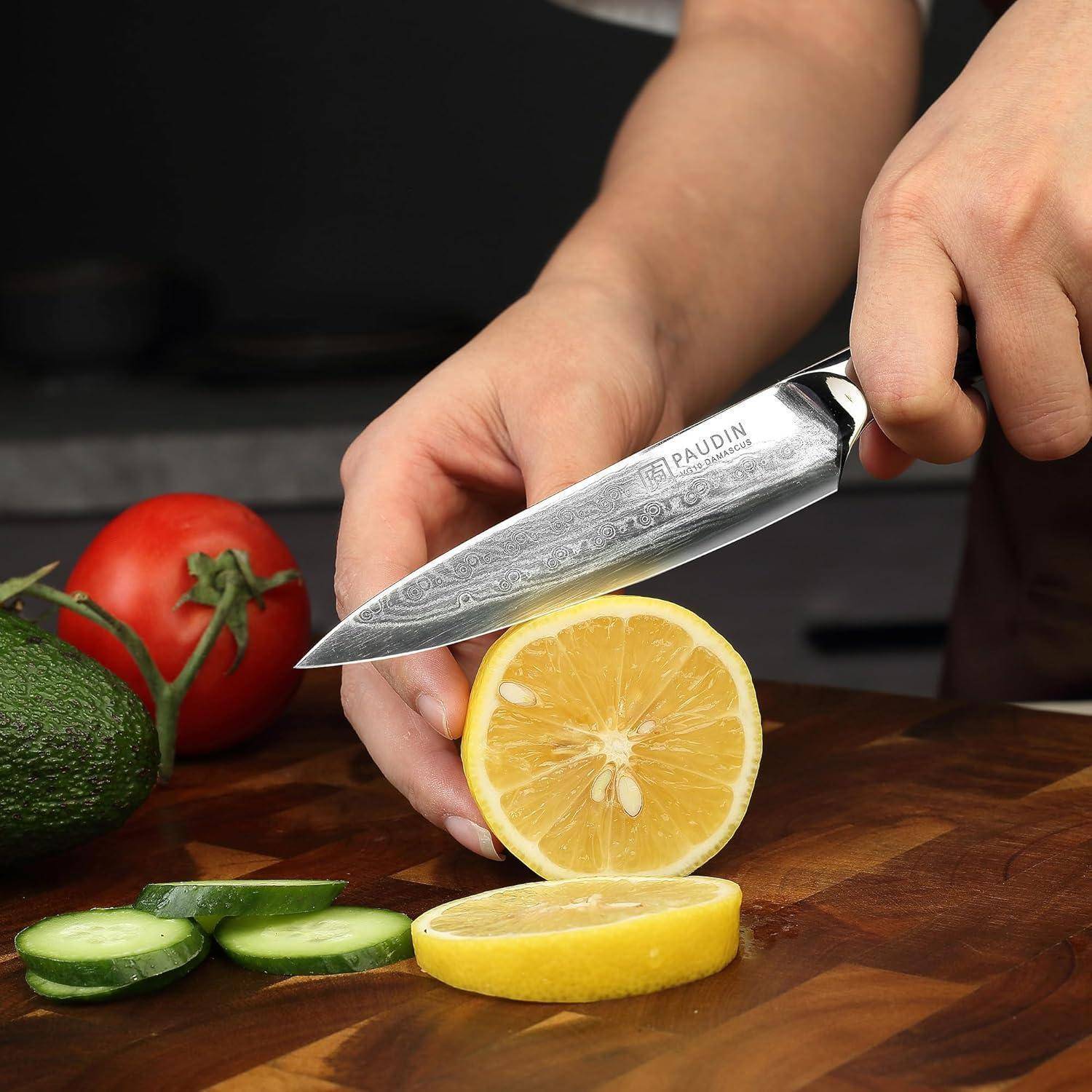 Eine Hand schneidet eine Zitrone auf einem Holzschneidebrett mit einem Messer. In der Nähe liegen Gurkenscheiben, eine Avocado und eine Tomate.