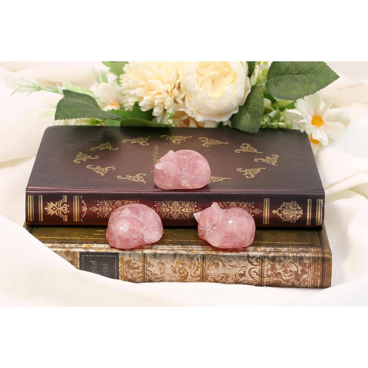 Drei Rosenquarz-Kristalle in Schweineform sitzen auf kunstvoll verzierten Büchern mit Blumenmuster, vor einem Blumenhintergrund.