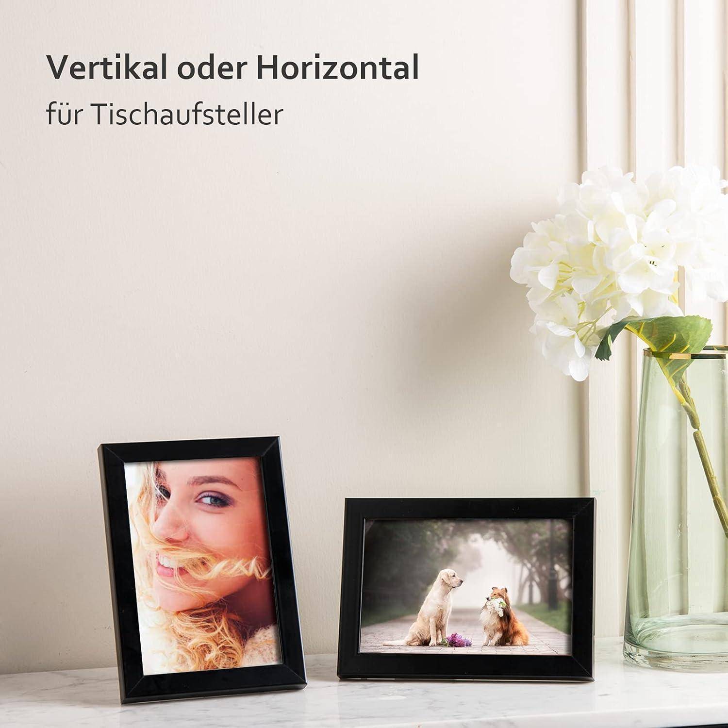 Zwei Bilderrahmen auf einem Tisch; einer hochkant mit einem Frauenporträt, einer quer mit zwei Tieren. Weiße Blumen in einer Vase rechts daneben.