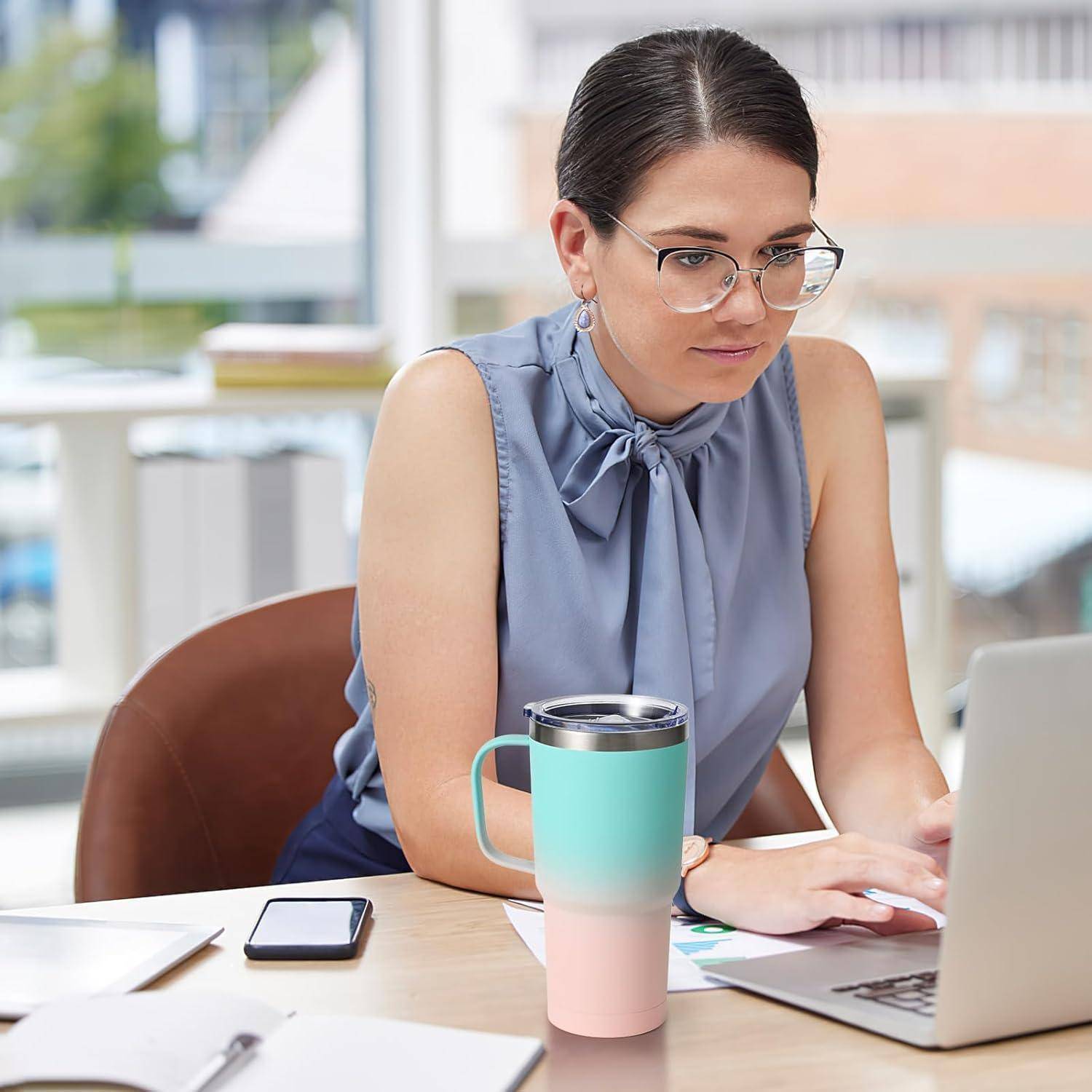 Eine Frau in einer blauen Bluse und mit Brille, die an einem Schreibtisch arbeitet und auf einen Laptop schaut. Ein Thermobecher, ein Smartphone und Papiere befinden sich in der Nähe.
