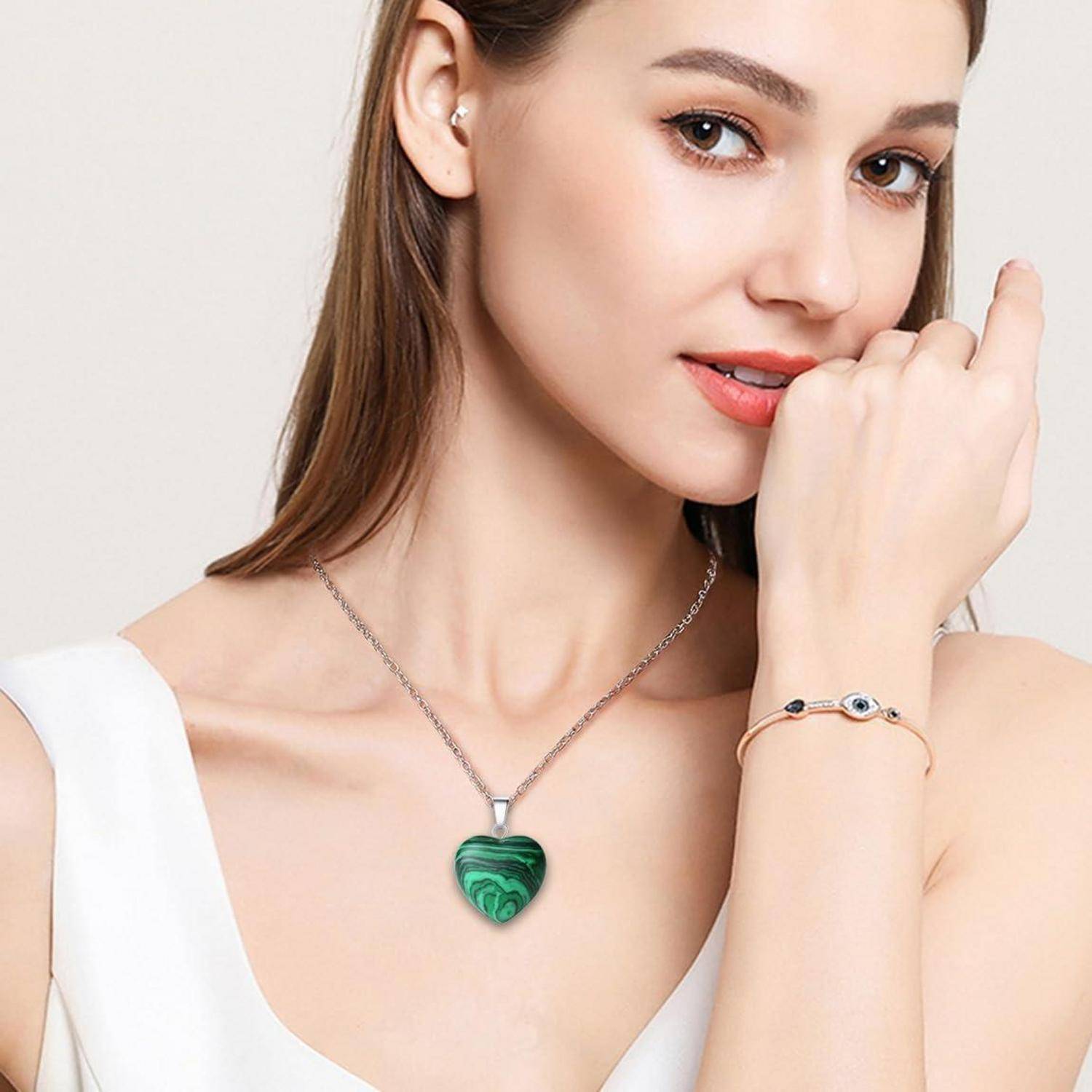 Eine Frau trägt einen grünen herzchenförmigen Anhänger und ein zartes Armband. Sie berührt ihr Kinn und präsentiert dabei die Schmuckstücke.