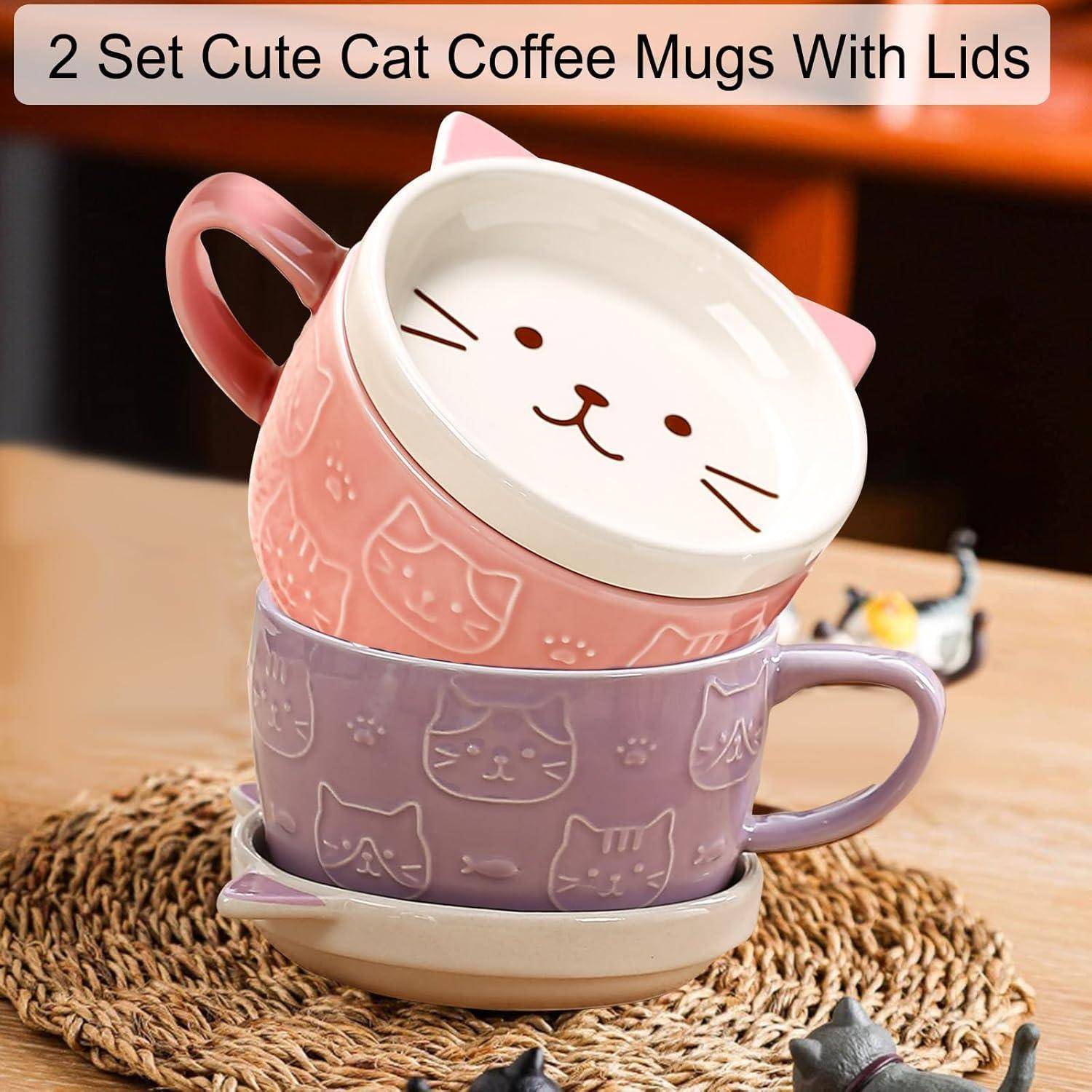 Zwei Kaffeebecher mit Katzen-Motiv und Deckel; einer rosa, einer lila, gestapelt auf einem Tisch.