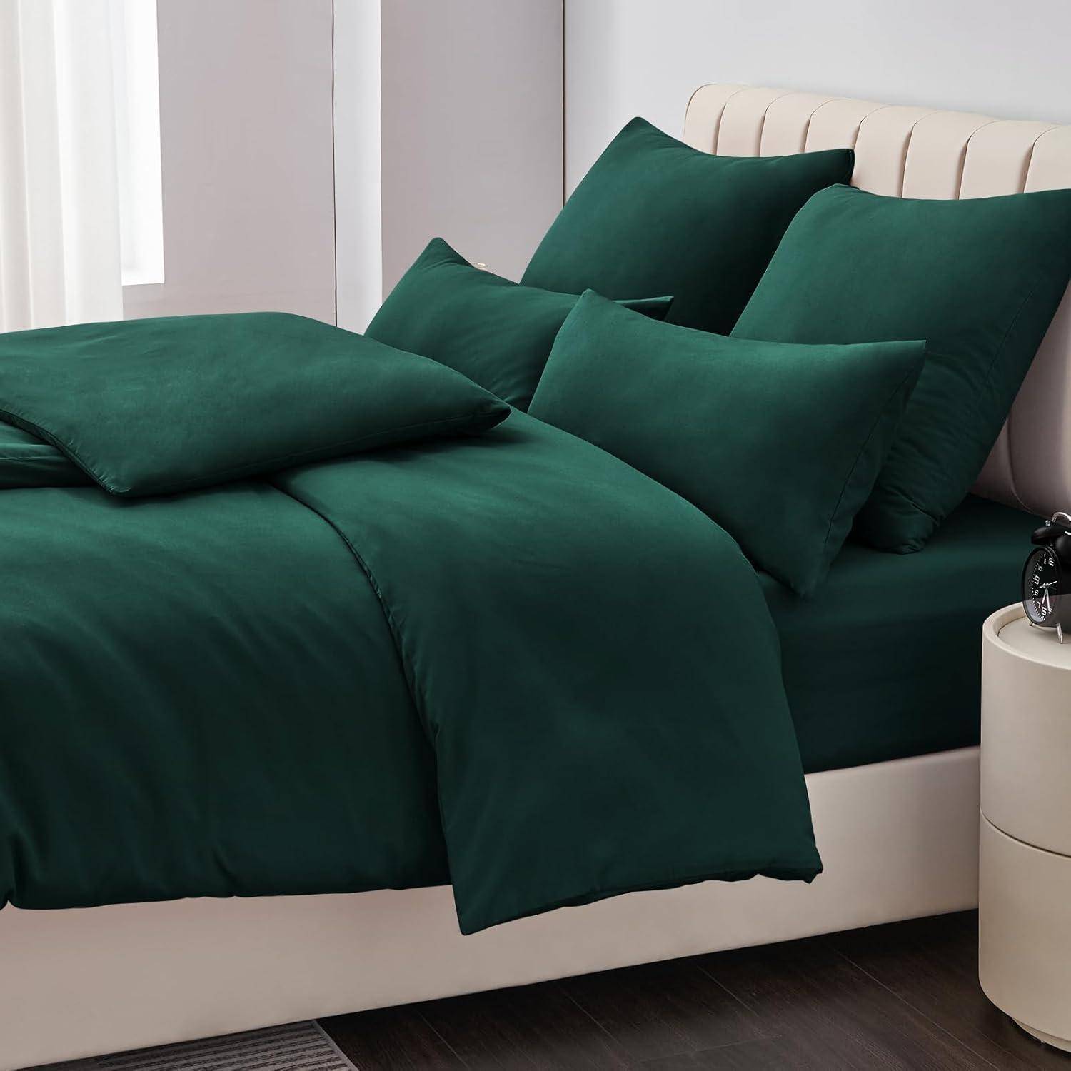 Ein sorgfältig hergerichtetes Bett mit dunkelgrüner Bettwäsche, bestehend aus Kissen, Bettdecke und Laken, vor einem hellgefärbten Kopfteil und Vorhängen.