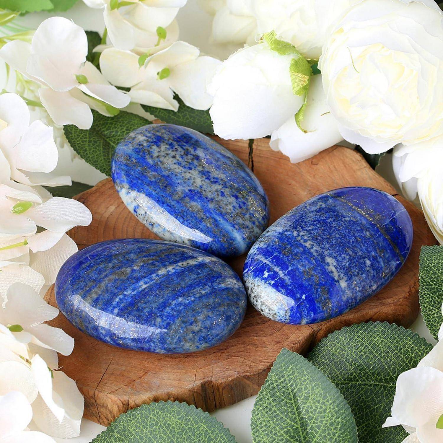 Drei geschliffene blaue Steine mit weißen Sprenkeln auf einer Holzoberfläche, umgeben von weißen Blumen und grünen Blättern.