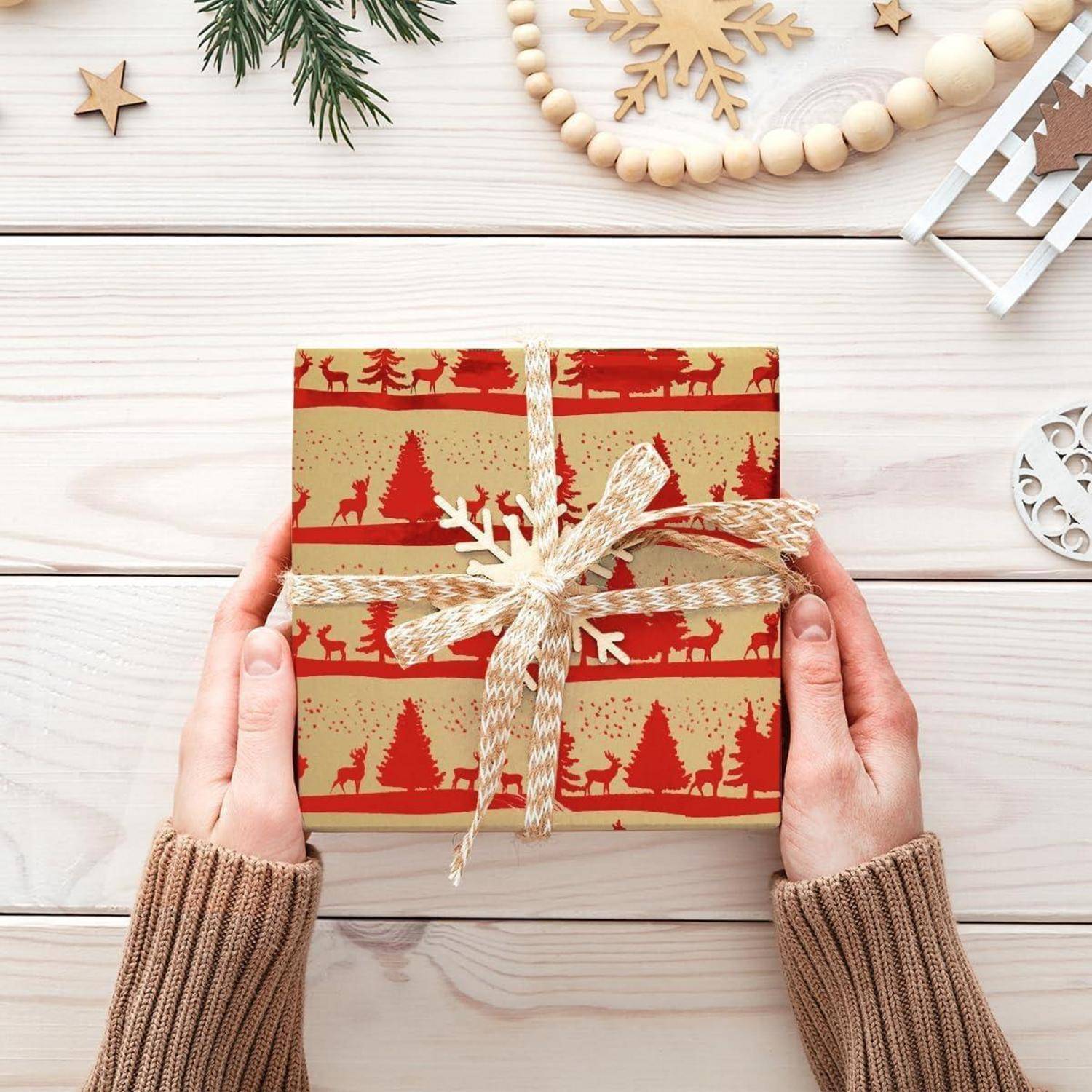 Hände, die ein festlich eingepacktes Geschenk mit roten Rentier- und Baummotiven halten, platziert auf einer Holzoberfläche, die mit Feiertagselementen dekoriert ist.