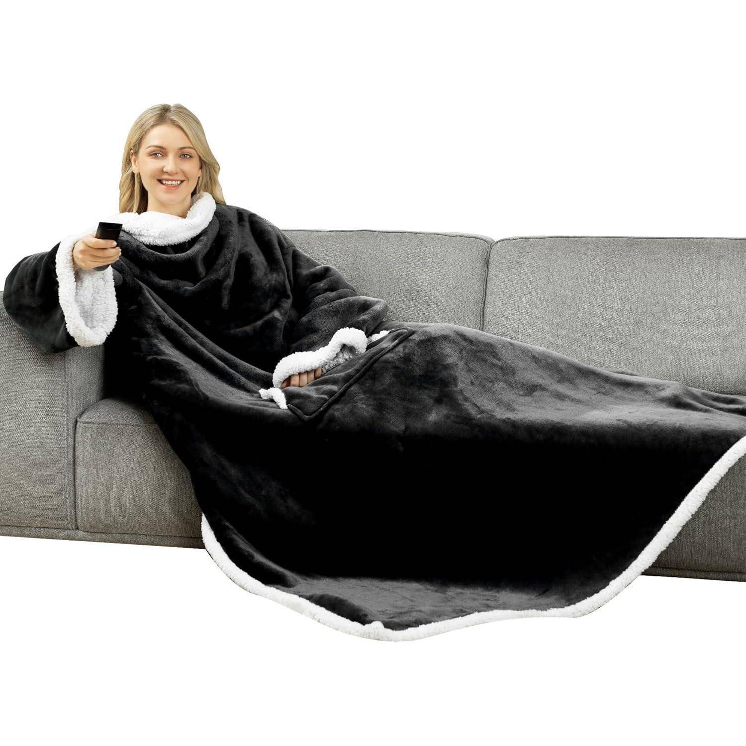 Eine Person auf einem grauen Sofa trägt eine große, schwarze Fleece-Decke mit weißer Einfassung und hält eine Fernbedienung, wirkt entspannt und behaglich.