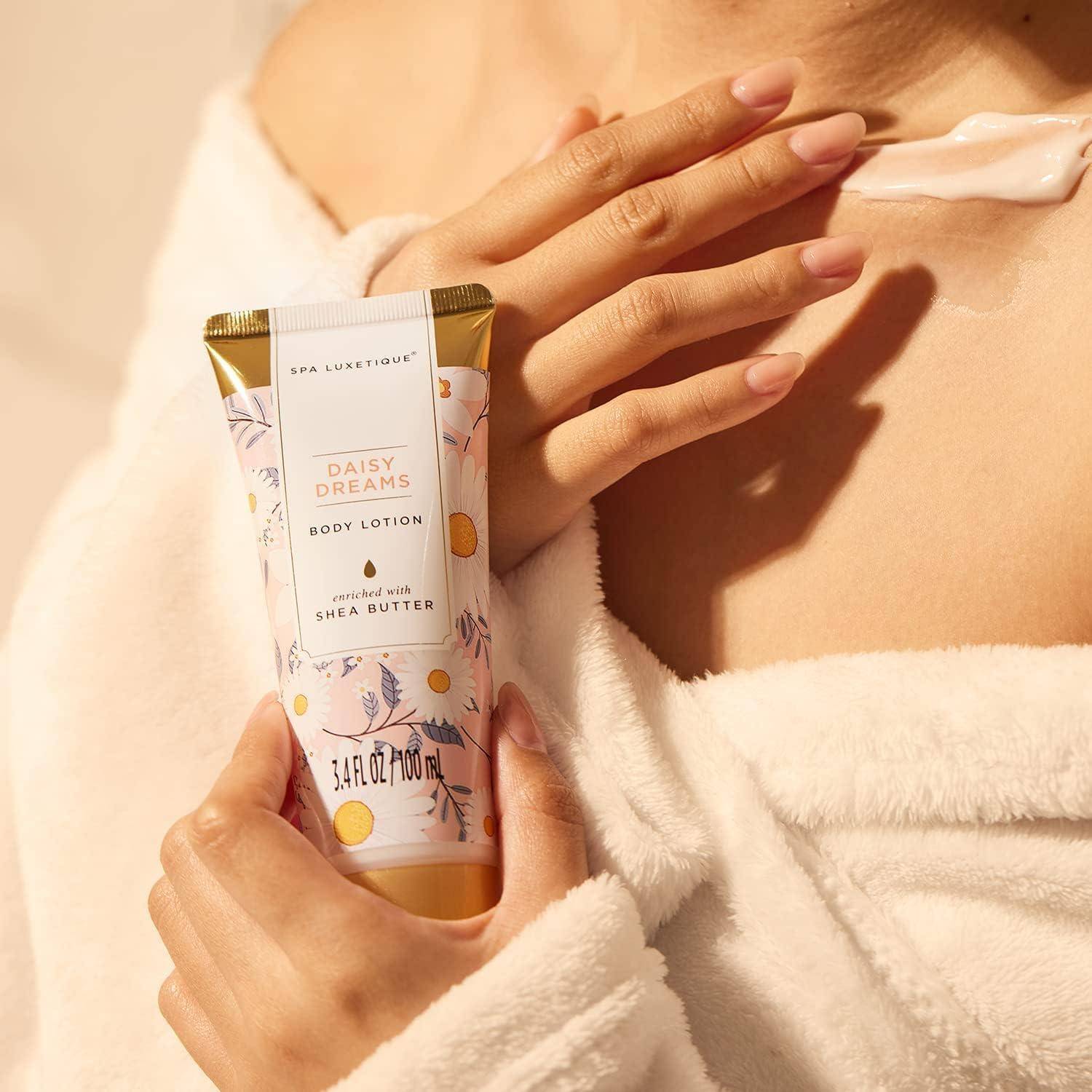 Eine Person trägt die Körperlotion ‚Daisy Dreams' mit Sheabutter auf ihre Schulter auf, während sie einen weißen Bademantel trägt, bei warmem, weichem Licht.