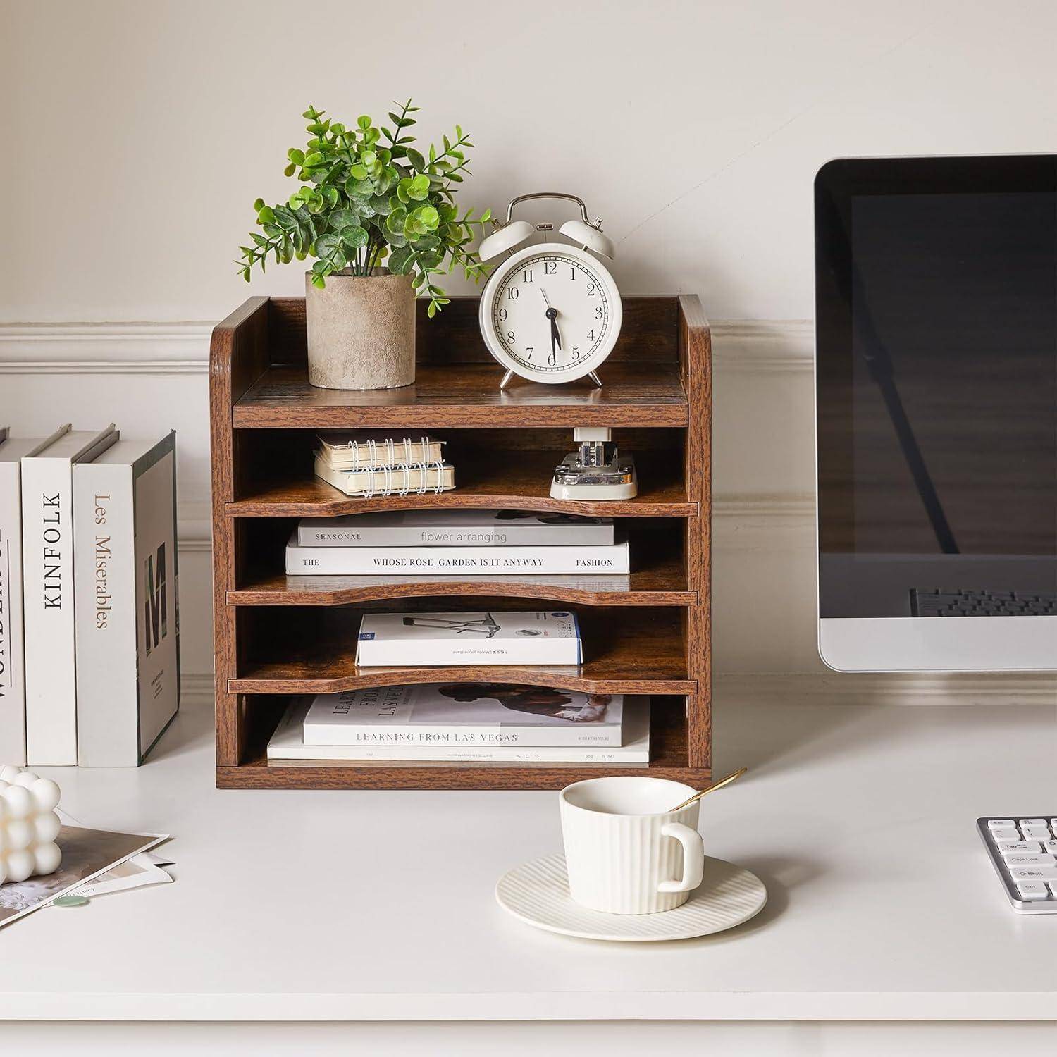Ein aufgeräumter Home-Office-Schreibtisch mit einem Holz-Organizer, auf dem Papiere und eine kleine Pflanze stehen, ein Wecker, Bücher auf der linken Seite und ein Computermonitor auf der rechten Seite.