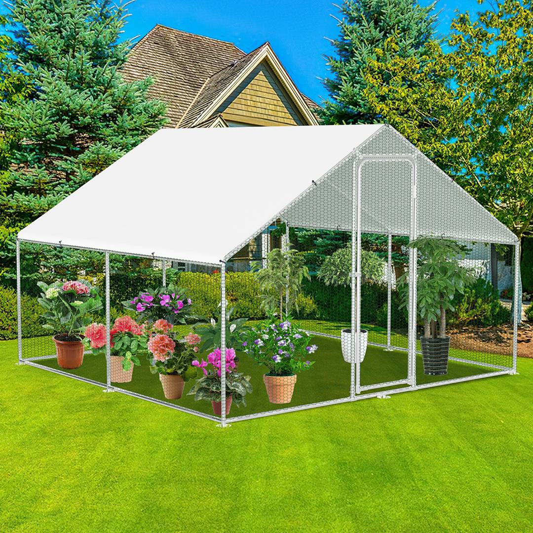 Ein weißer, metallgerahmter Gewächshaus auf Gras, in dem verschiedene Topfpflanzen und Blumen stehen. Eine Schaufel hängt an der Konstruktion. Im Hintergrund befinden sich hohe Bäume und ein Haus.